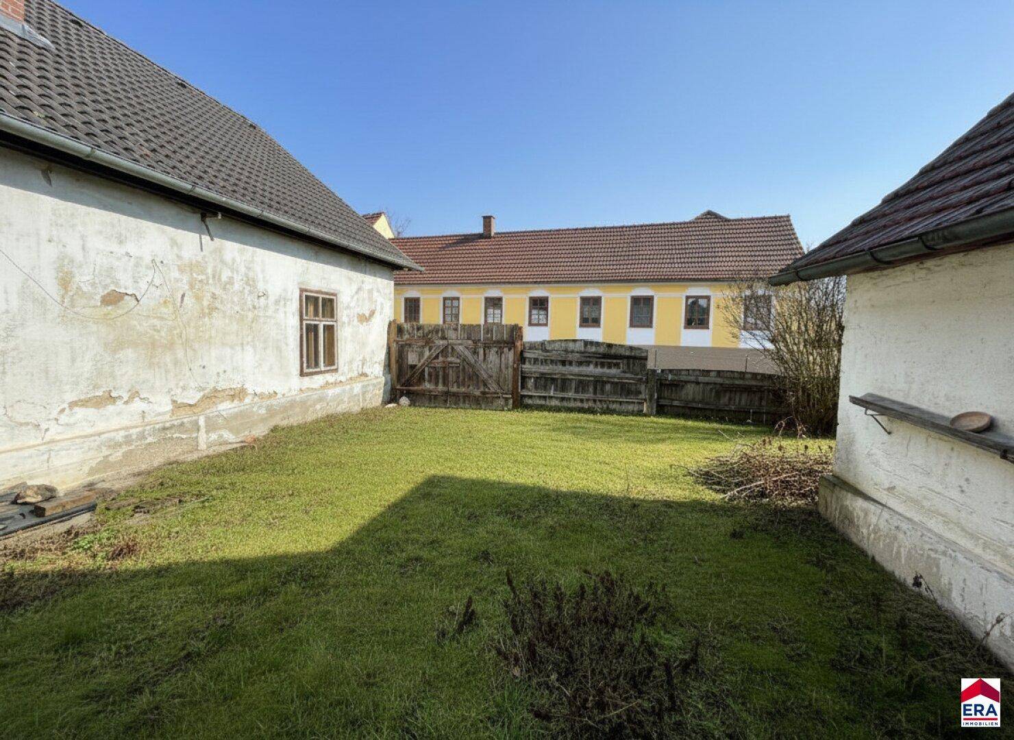 Gartenbereich zwischen zwei Gebäuden mit grünem Rasen und einem Holzzaun im Hintergrund.