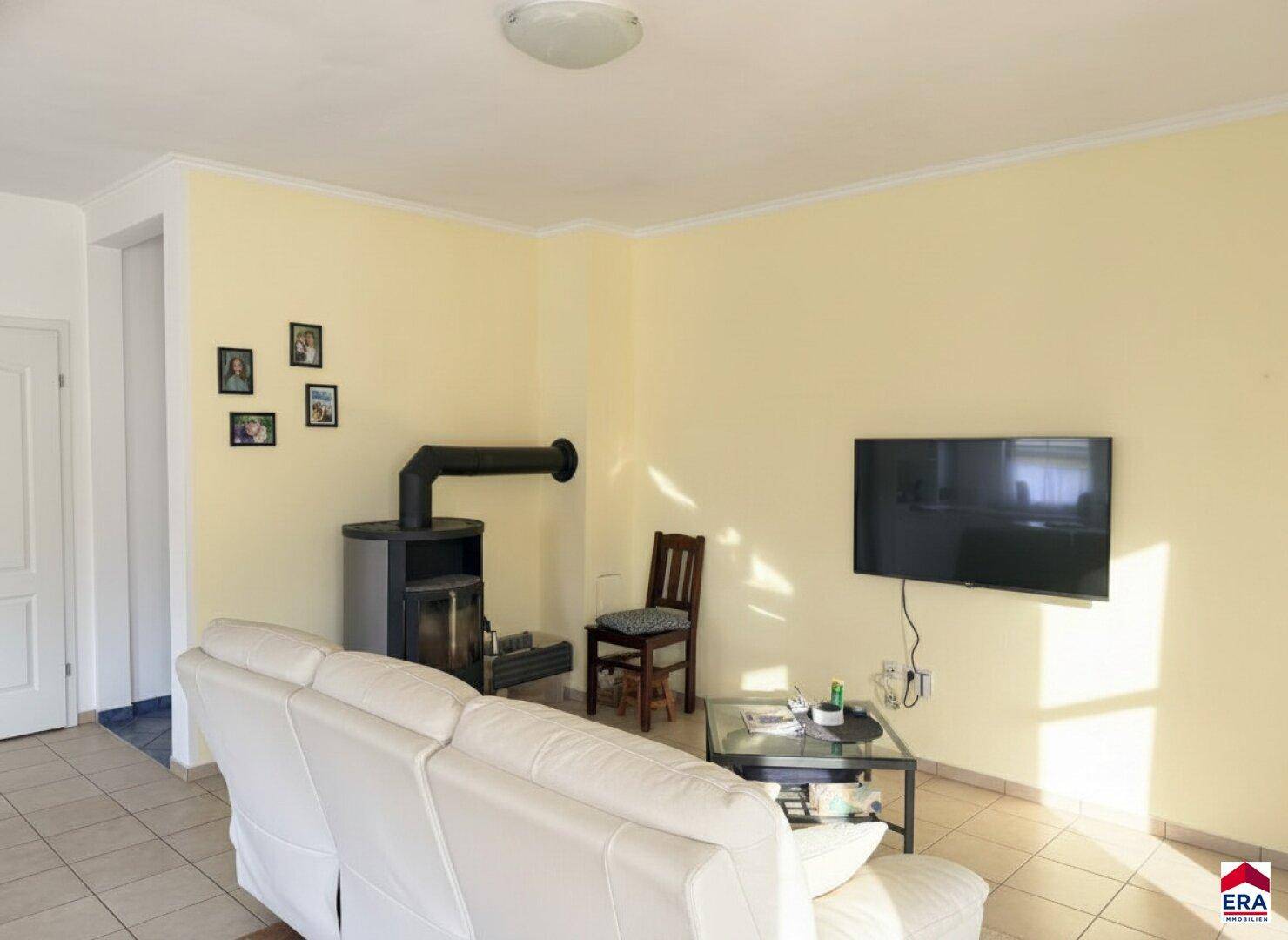 Helles Wohnzimmer mit Kaminofen, Fliesenboden und gemütlicher Sitzgelegenheit.