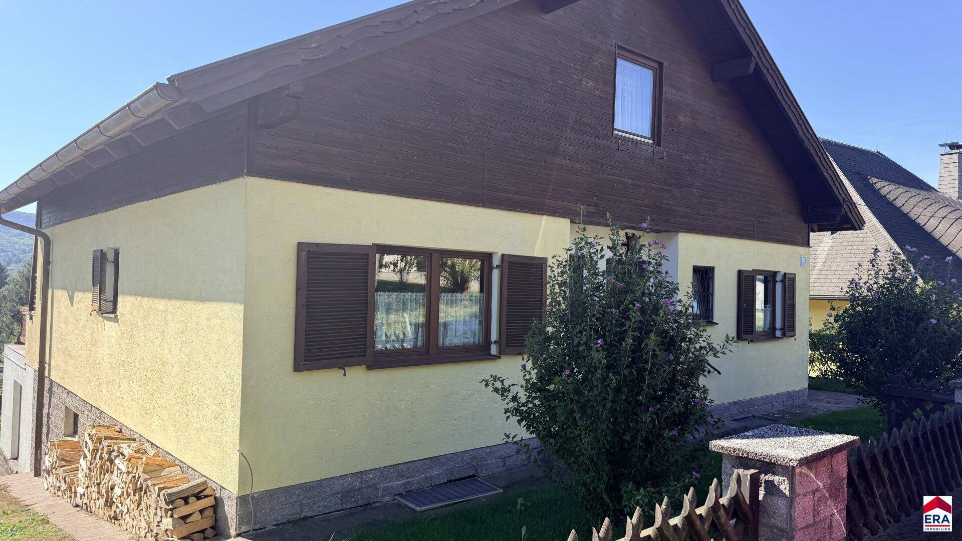 Seitenansicht des Hauses mit Holzfassade, Fensterläden und gestapeltem Brennholz.