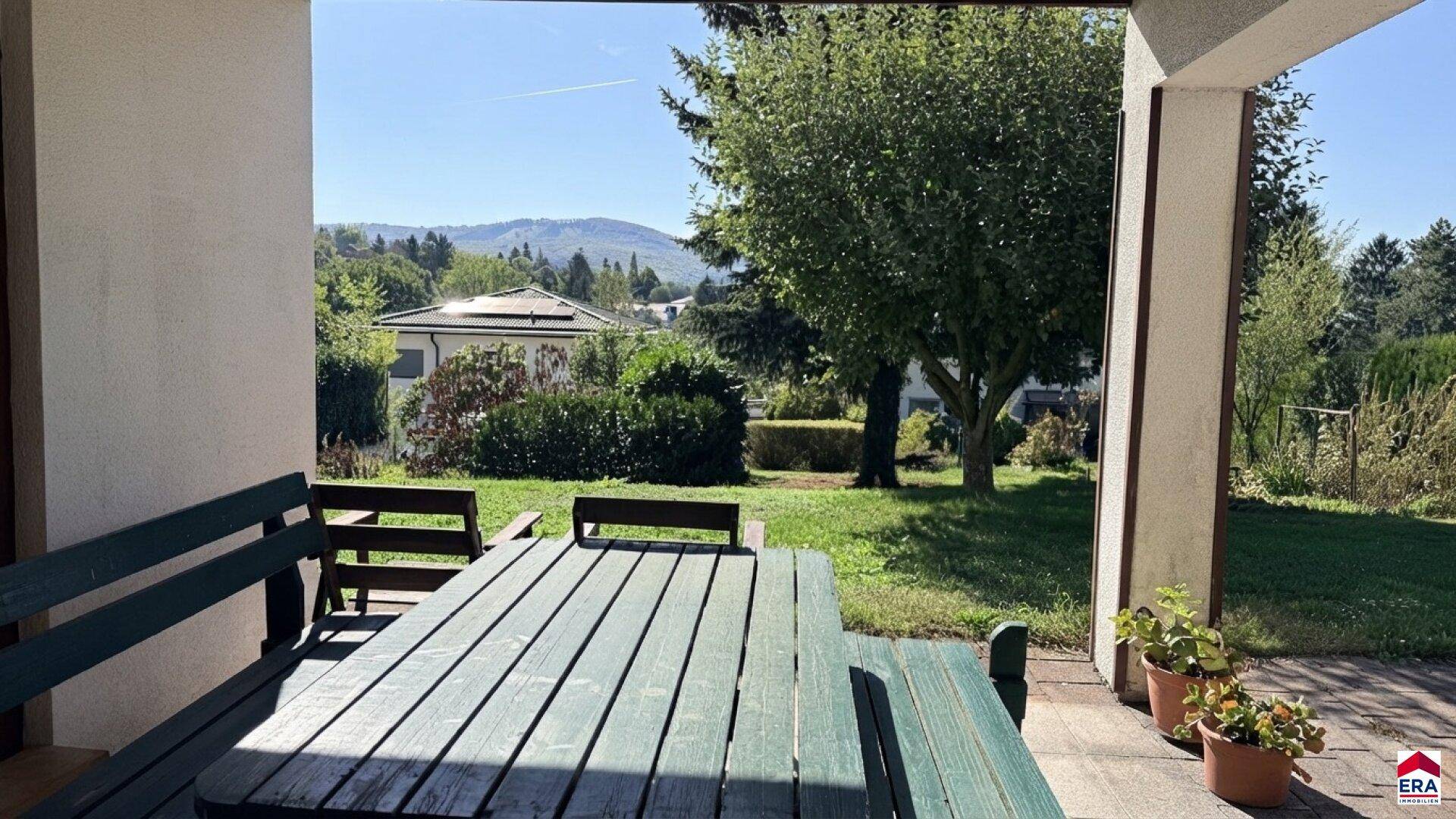 Überdachte Terrasse mit Holztisch und Bänken, umgeben von Grünflächen.