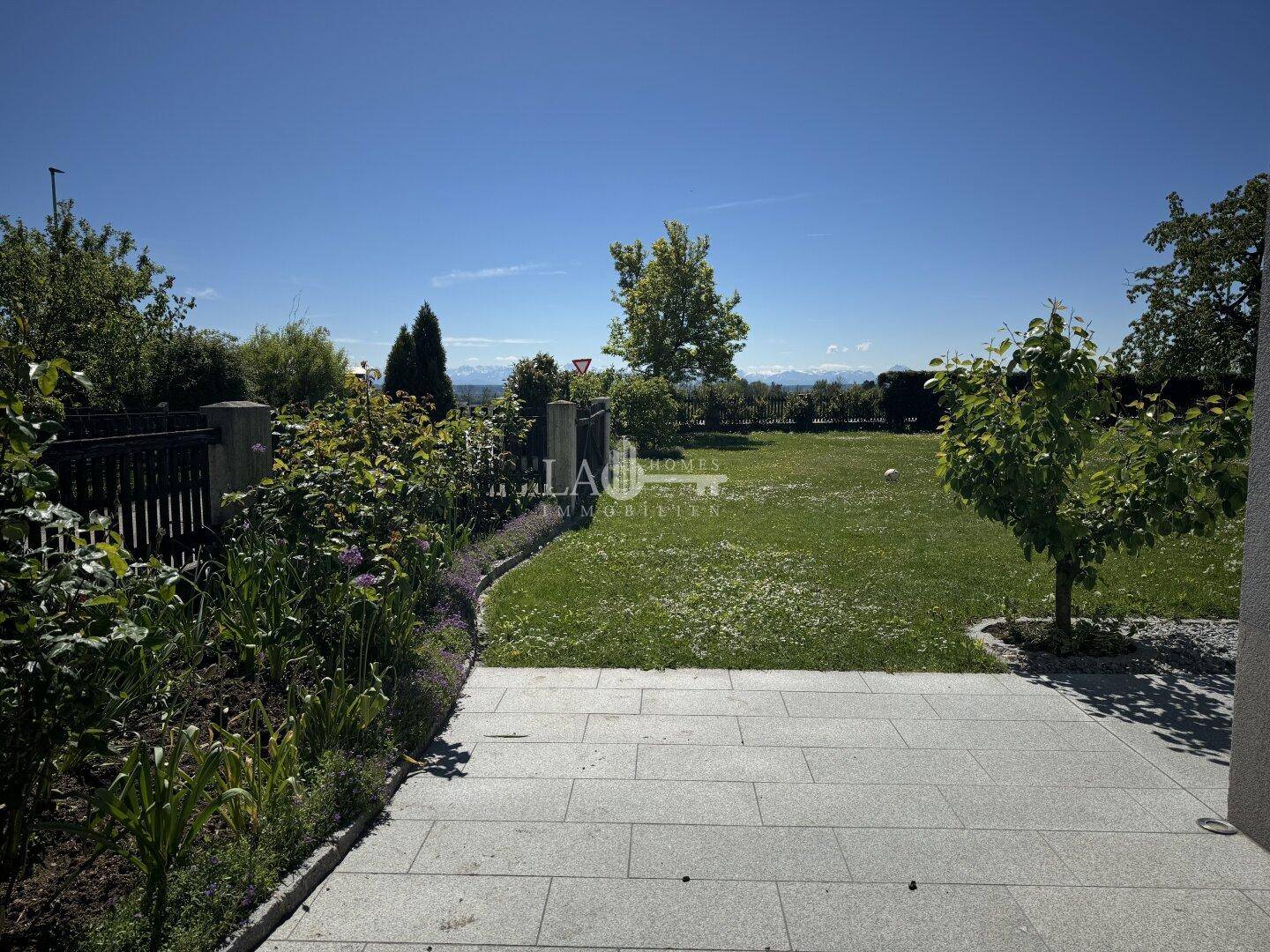 Gepflegter Gartenbereich mit Blumenbeeten, Steinfliesen und weitem Blick ins Grüne.