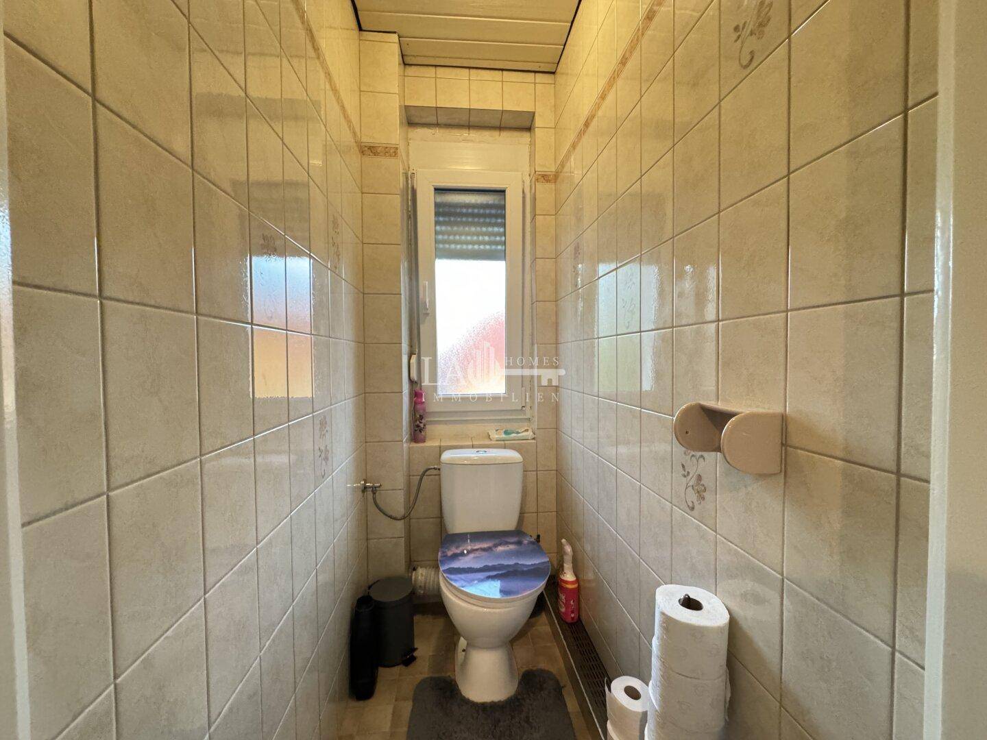 Separates WC mit Fliesen, wandhängendem WC und kleinem Fenster.