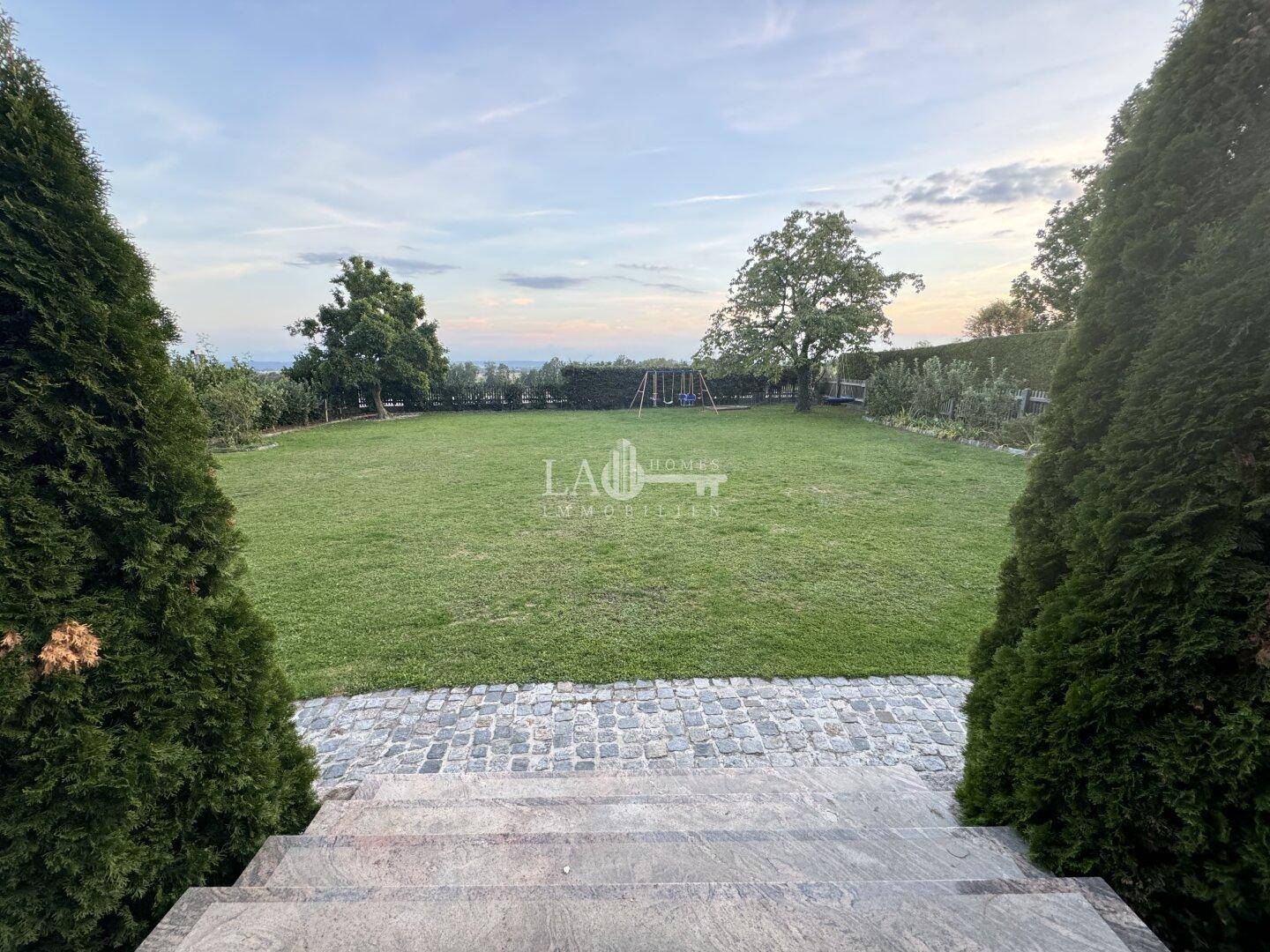 Weitläufiger Garten mit Rasenfläche, Spielplatz im Hintergrund und Steintreppe.