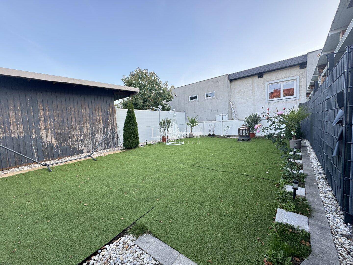 Gepflegter Garten mit Kunstrasenfläche, Gartenhaus und dekorativen Pflanzen.