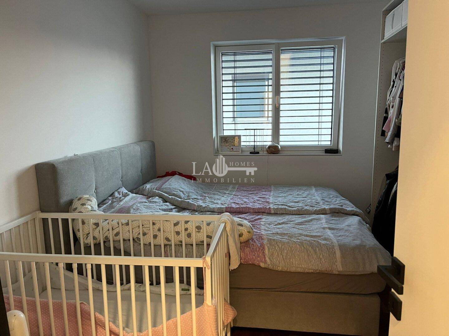 Helles Schlafzimmer mit Doppelbett, Babybett und Fenster mit Jalousien.