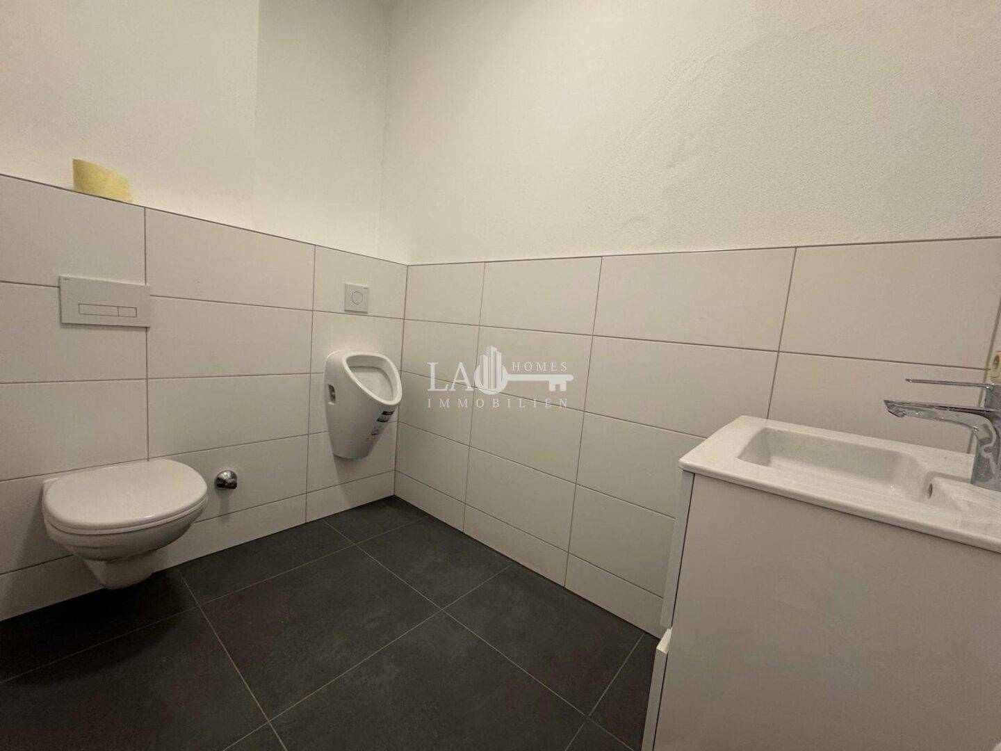 Modernes Gäste-WC mit dunklen Fliesen, wandmontierter Toilette und Urinal, in einem gepflegten Zustand.