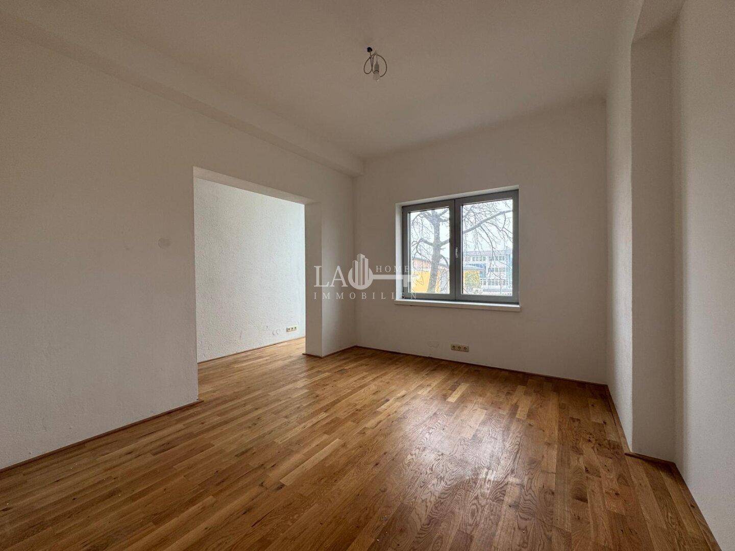 Leeres Zimmer mit Holzboden und einem Fenster, das viel natürliches Licht hereinlässt und einen Blick nach draußen bietet.