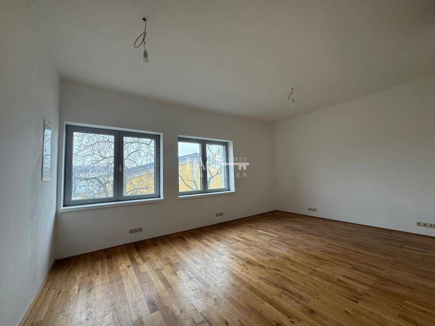 Helles Schlafzimmer mit Holzboden und zwei Fenstern, die einen Blick auf die Bäume und die Umgebung bieten.