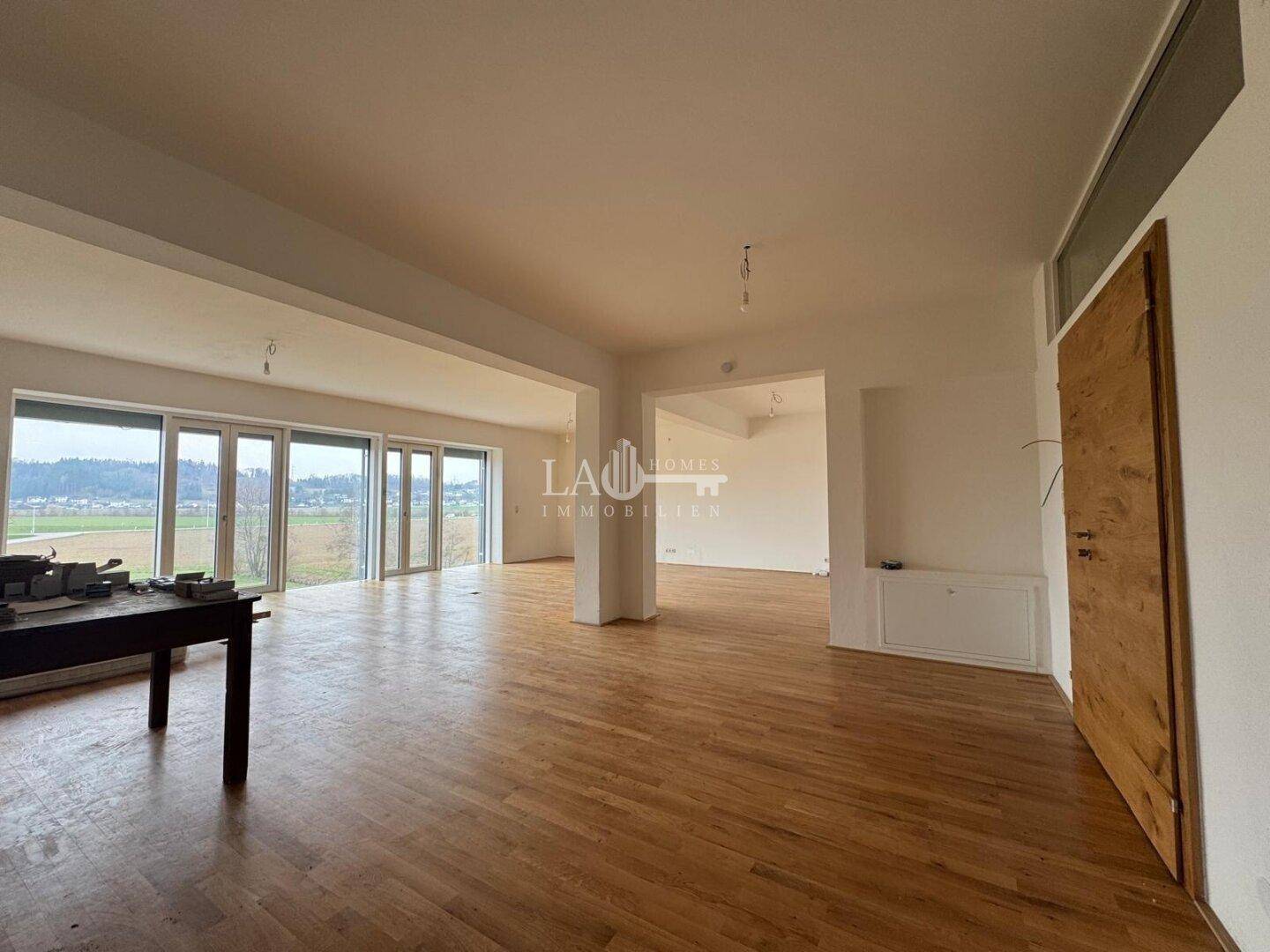 Heller Wohnbereich mit Holzboden und breiter Fensterfront, die einen weiten Blick auf die umliegende Landschaft ermöglicht.