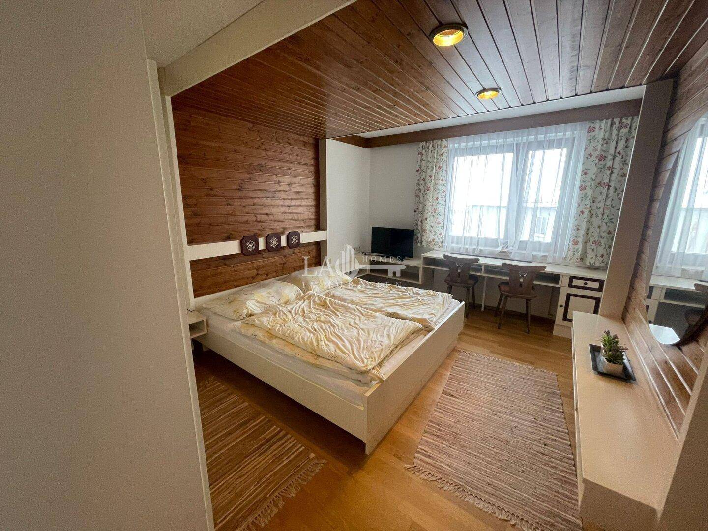 Gemütliches Schlafzimmer mit Holzwänden, Doppelbett und Schreibtischbereich am Fenster.