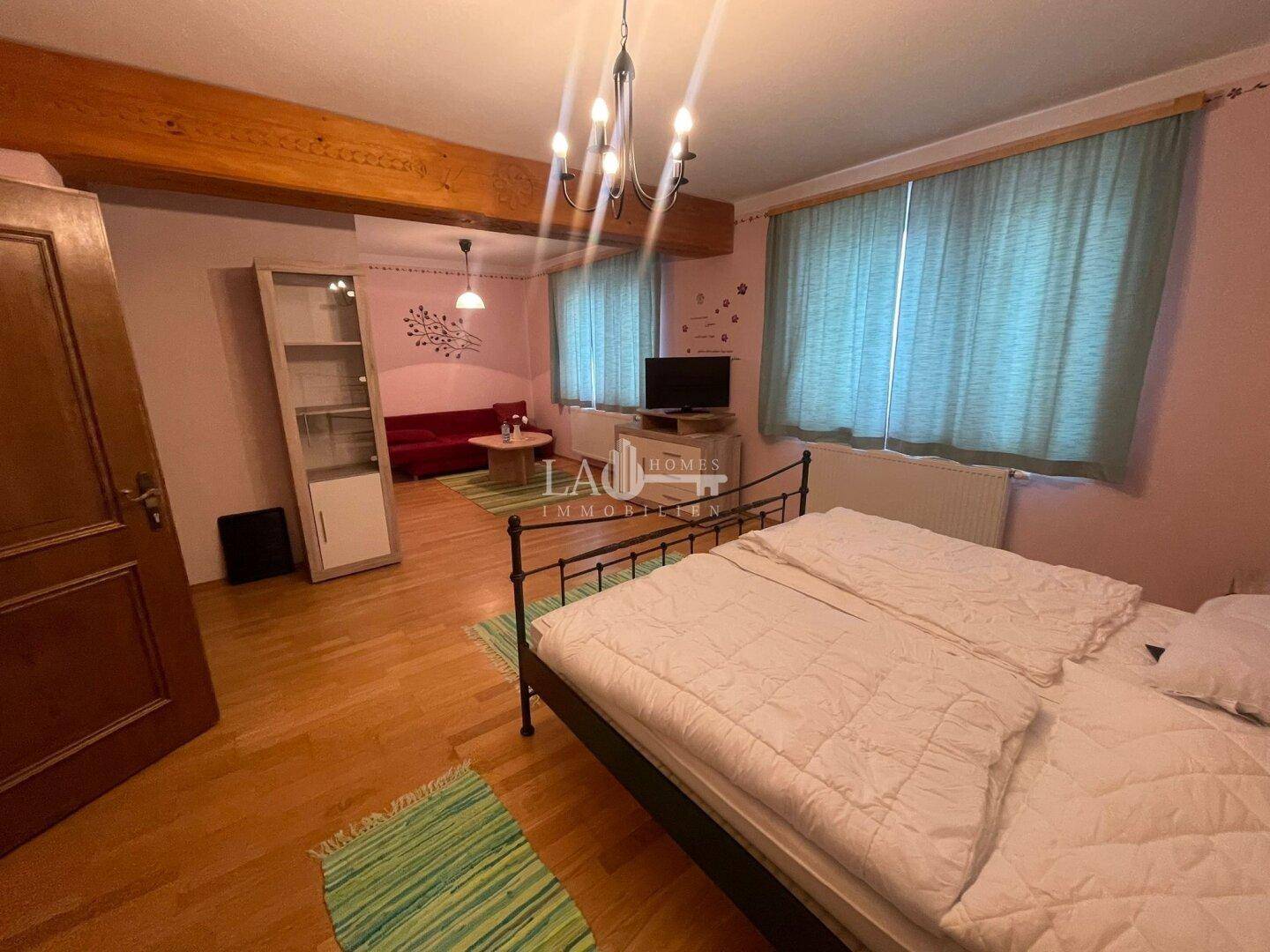 Schlafzimmer mit Holzboden, Doppelbett und einem separaten Wohnbereich mit Sofa im Hintergrund.