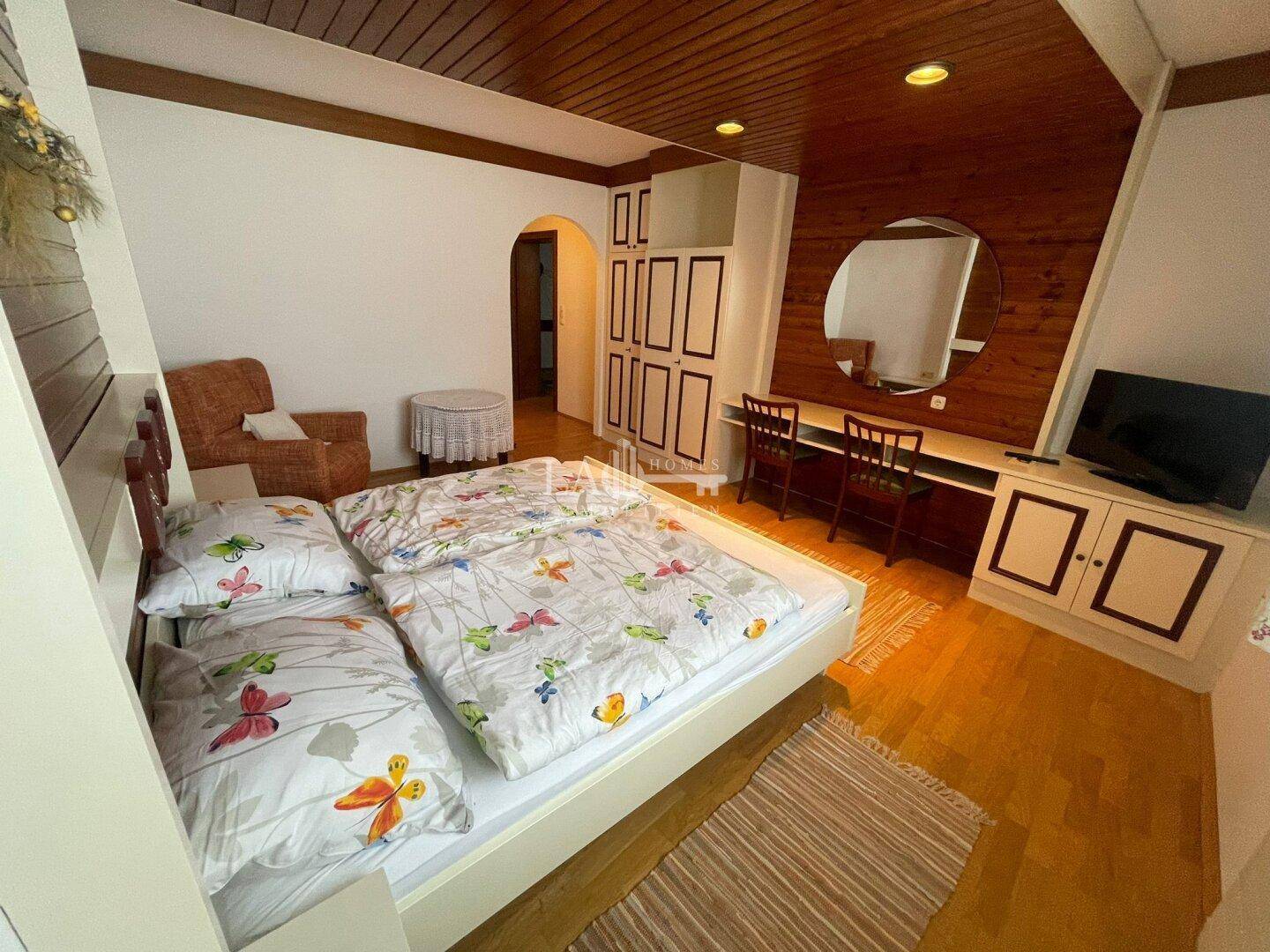 Schlafzimmer mit Holzboden, Doppelbett, Schminktisch und einem Sessel in warmer Atmosphäre.