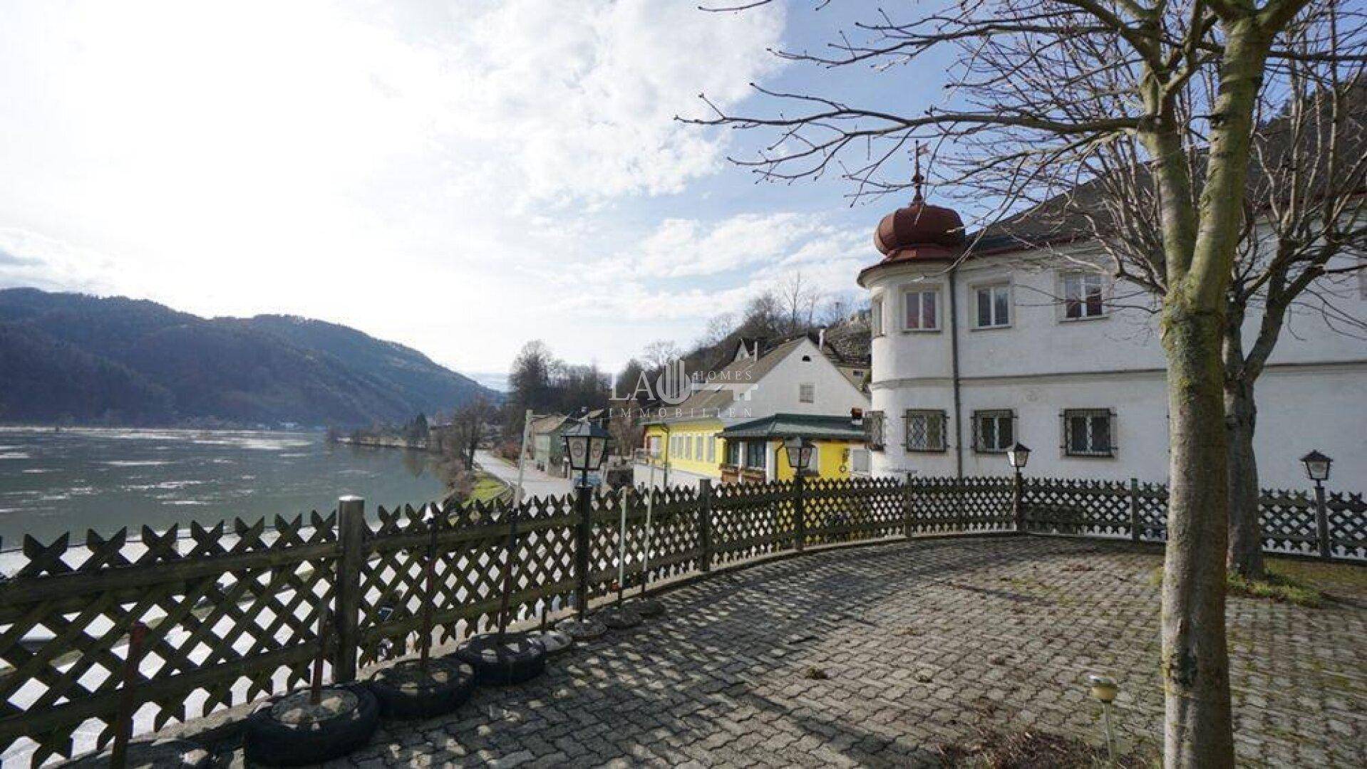 Gepflasterter Hofbereich mit Holzzaun und Blick auf den Fluss sowie die Berge, neben einem historischen Gebäude.