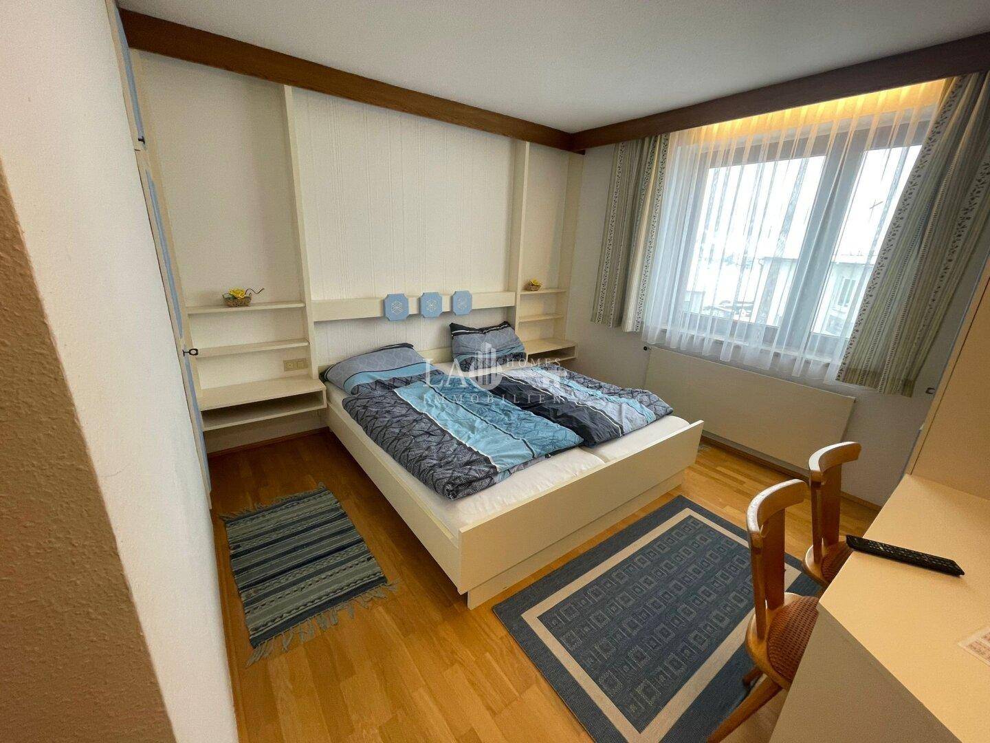 Helles Schlafzimmer mit Doppelbett, Holzbalken und einem Schreibtischbereich am Fenster.