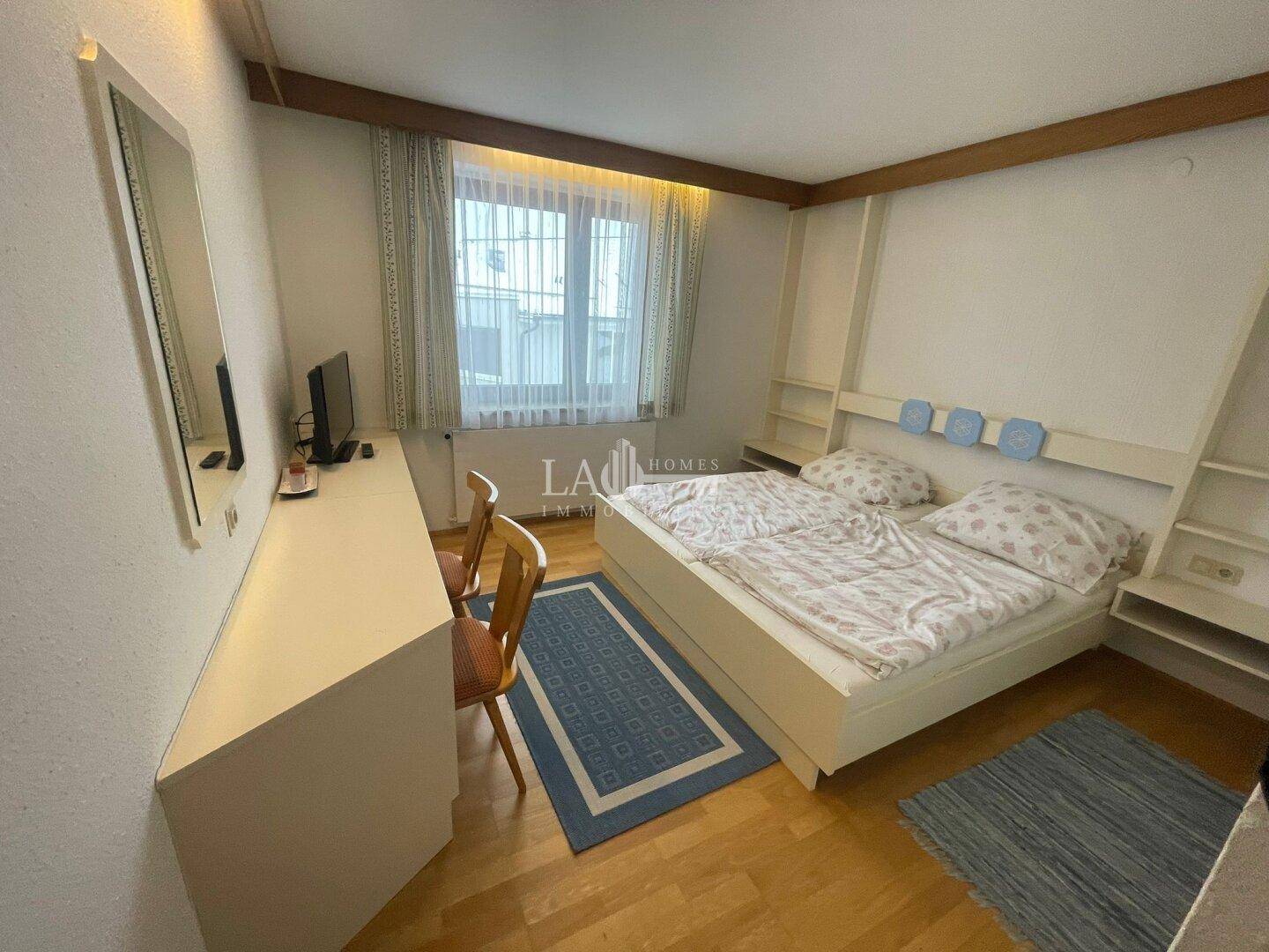 Schlafzimmer mit Doppelbett, Holzboden und einem langen Schreibtisch unter dem Fenster.