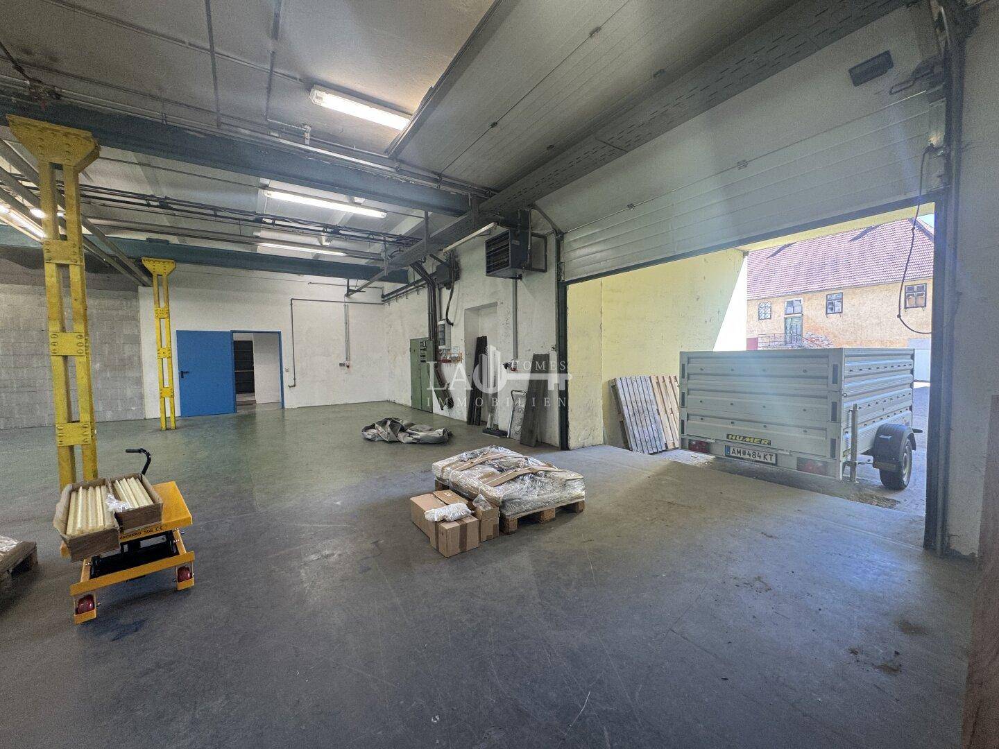 Große, leere Lagerhalle mit Betonboden, hohen Decken und einem geöffneten Rolltor zur Außenseite.