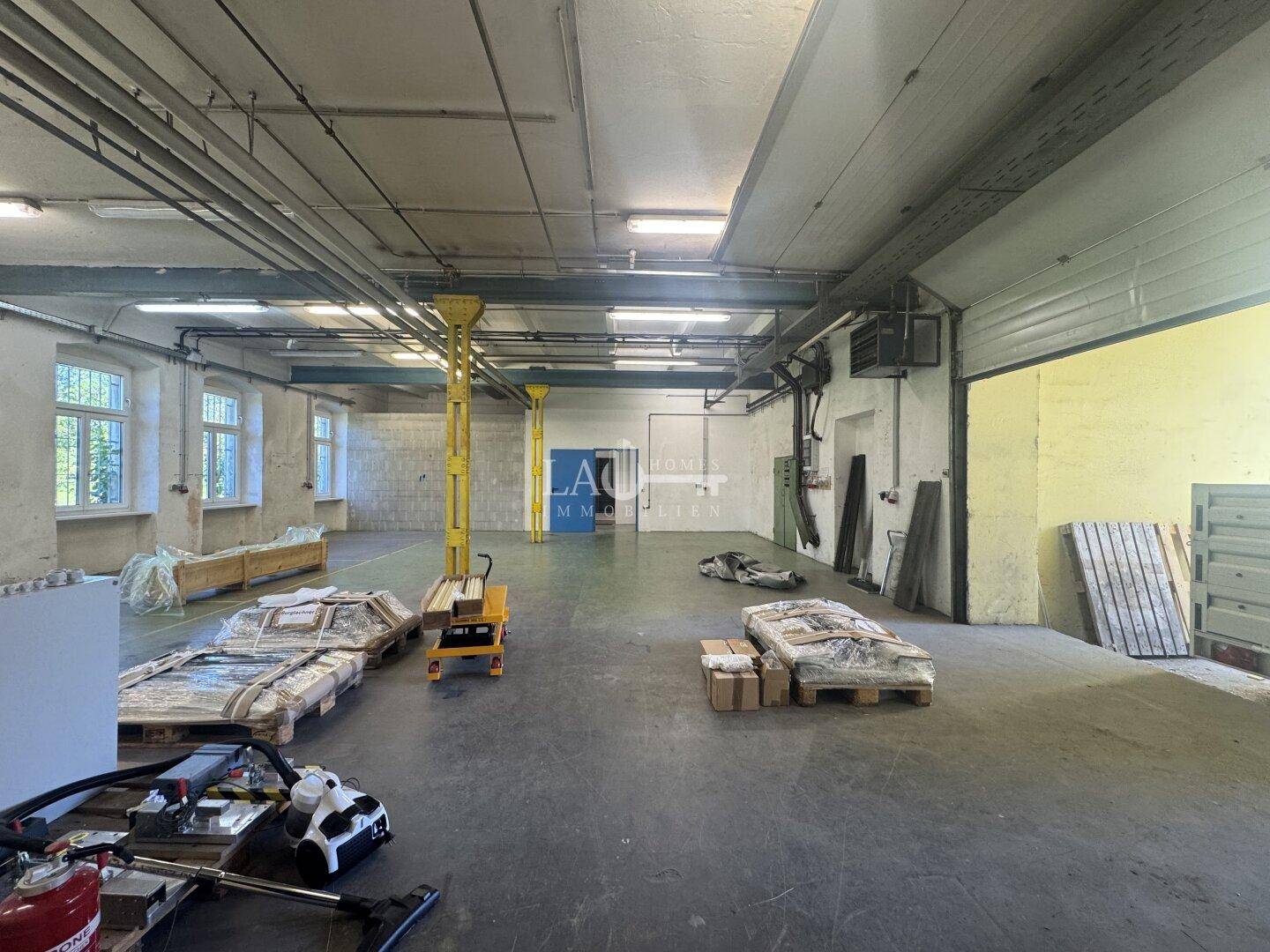 Geräumige Lagerhalle mit Betonboden, hohen Decken und mehreren Fenstern, die Tageslicht hereinlassen.