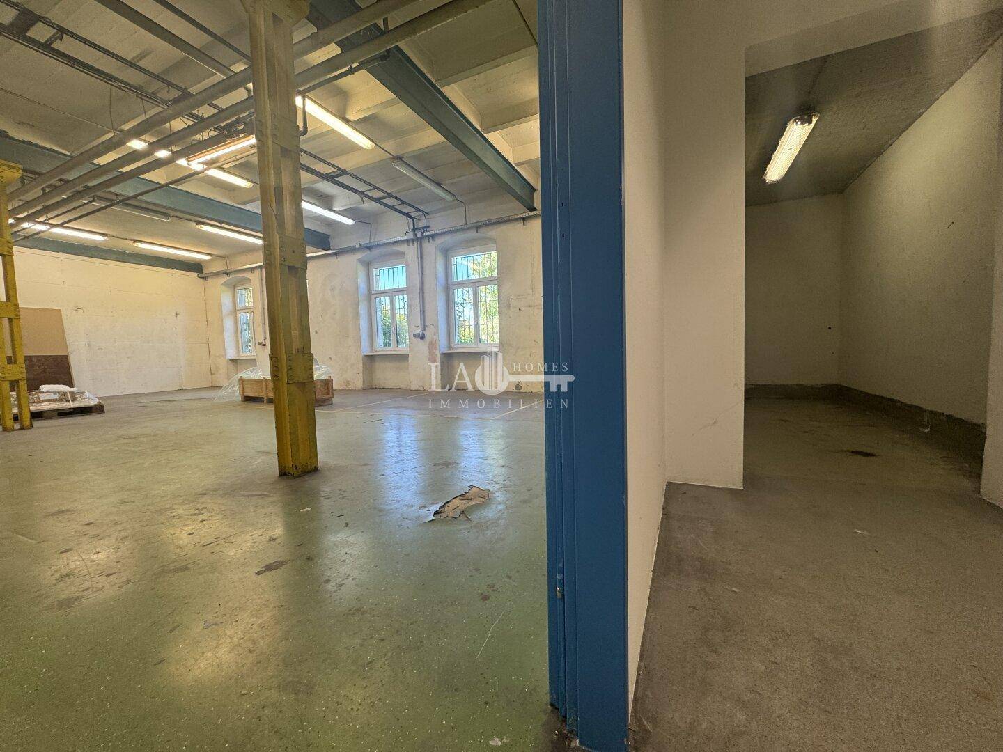 Blick in die Lagerhalle mit grünem Betonboden und einem blauen Türrahmen, der zu einem Nebenraum führt.