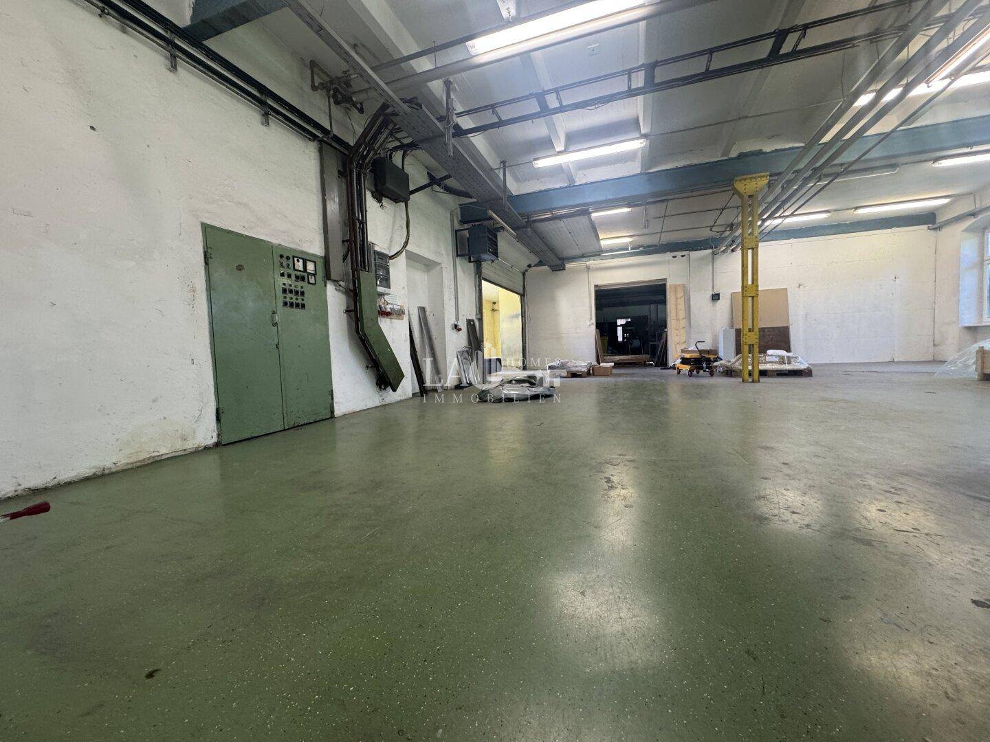 Industrielle Lagerhalle mit grünem Betonboden, sichtbaren Installationen und einem geöffneten Tor im Hintergrund.