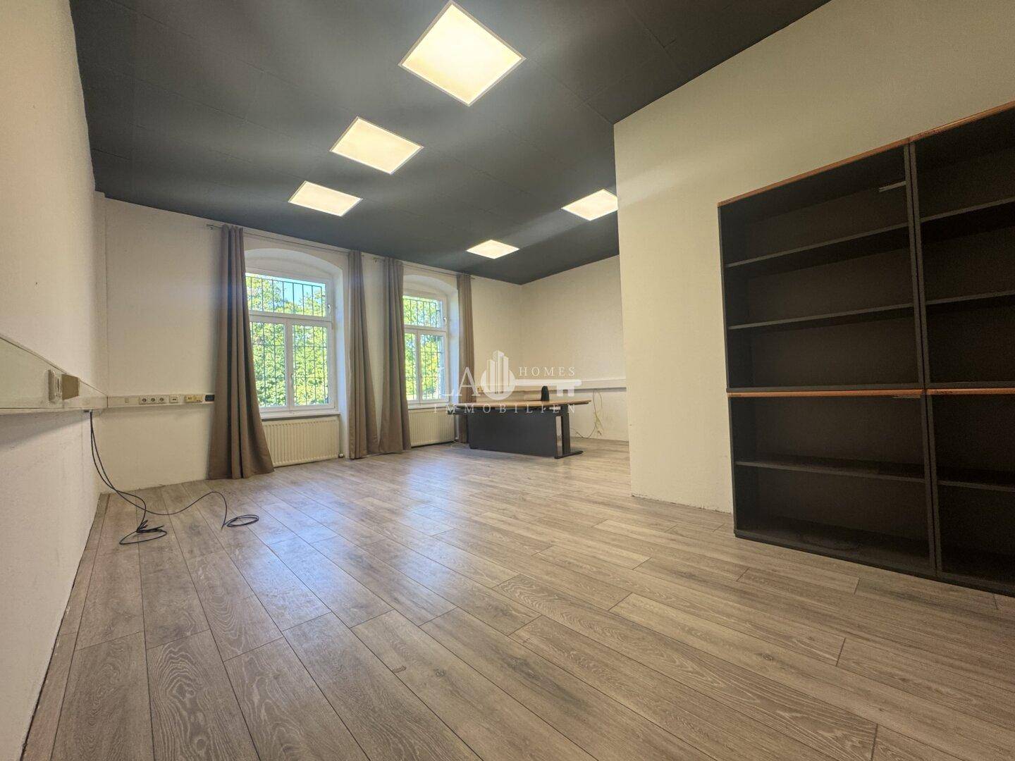 Modernes Büro mit Laminatboden, weißen Wänden, dunkler Decke und großen Fenstern mit Blick ins Grüne.