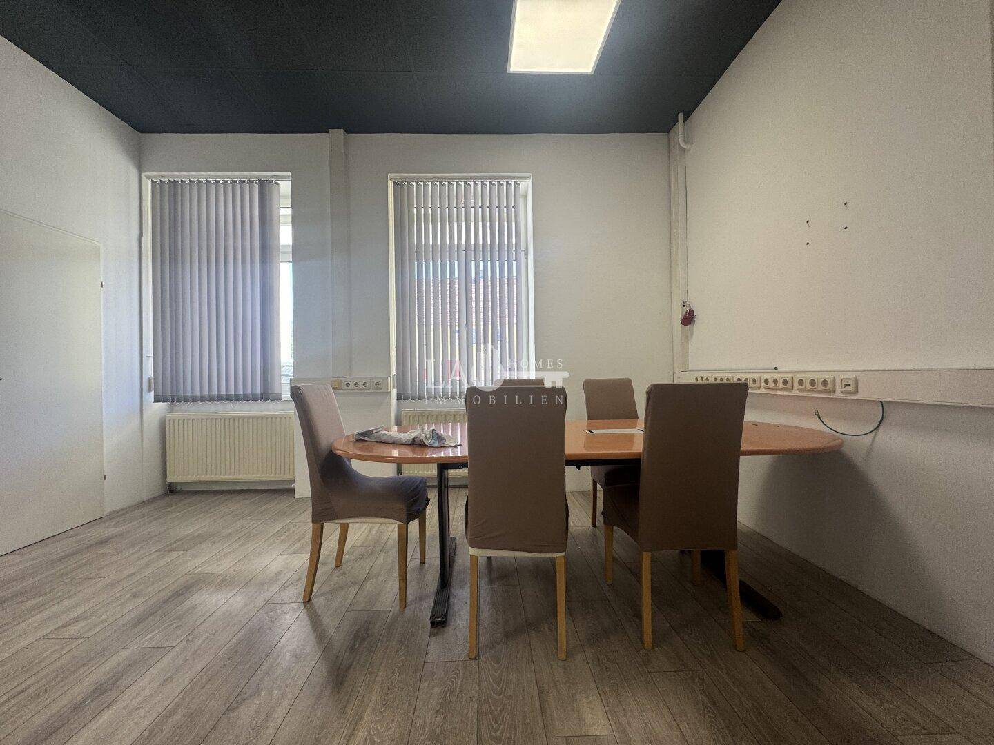 Konferenzraum im Büro mit Laminatboden, einem ovalen Tisch und Stühlen sowie Jalousien an den Fenstern.