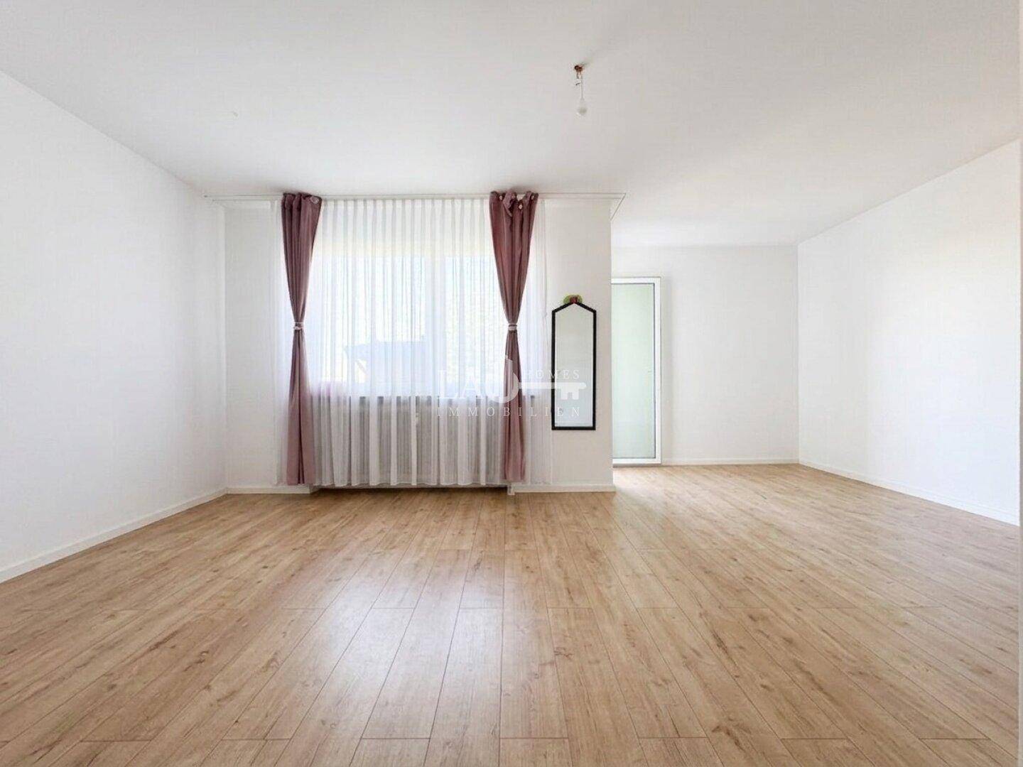 Geräumiges, leeres Wohnzimmer mit hellem Laminatboden und großem Fenster.