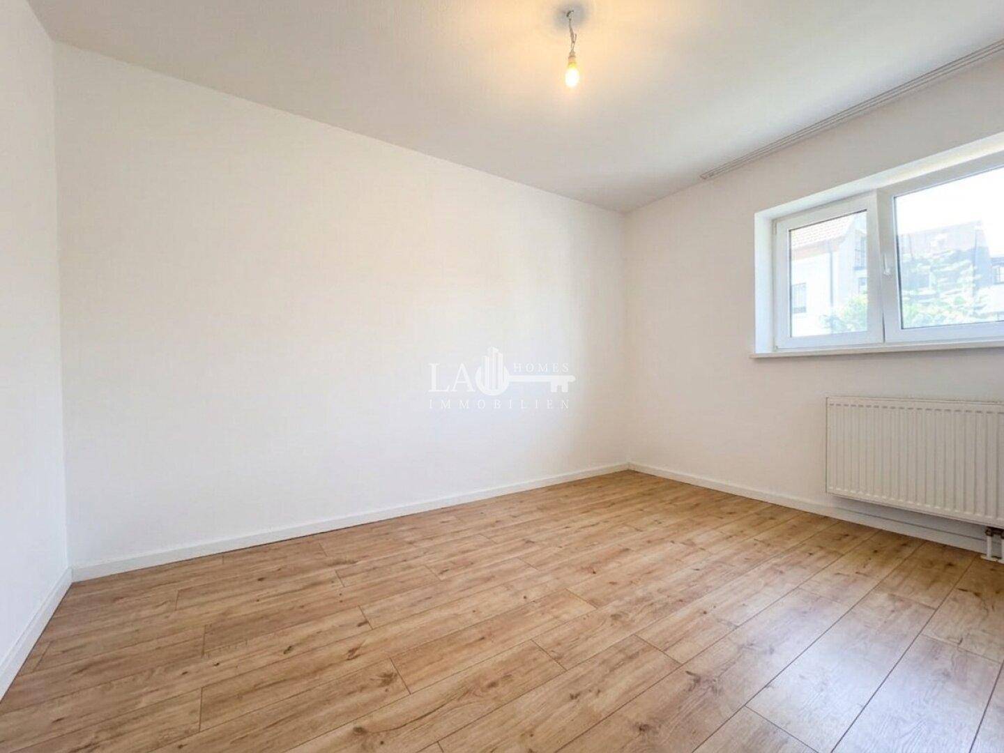 Leeres Schlafzimmer mit hellem Laminatboden, weißen Wänden und Fenster mit Heizkörper.