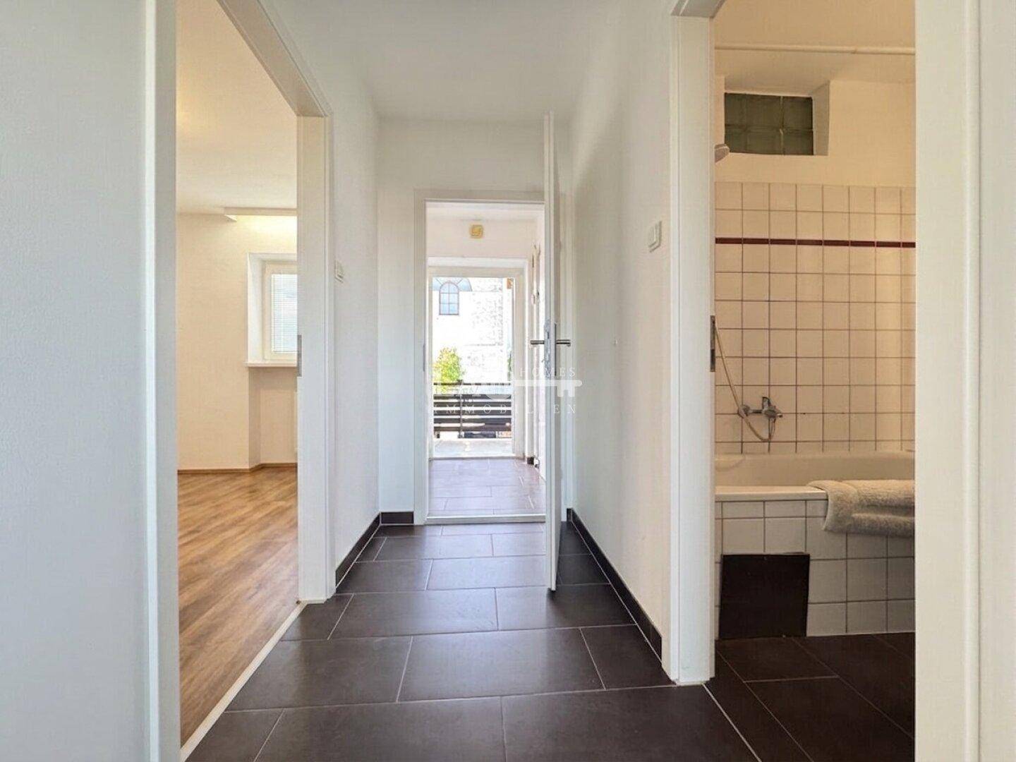 Flur mit dunklen Fliesen, Blick ins Badezimmer und auf einen Balkon.