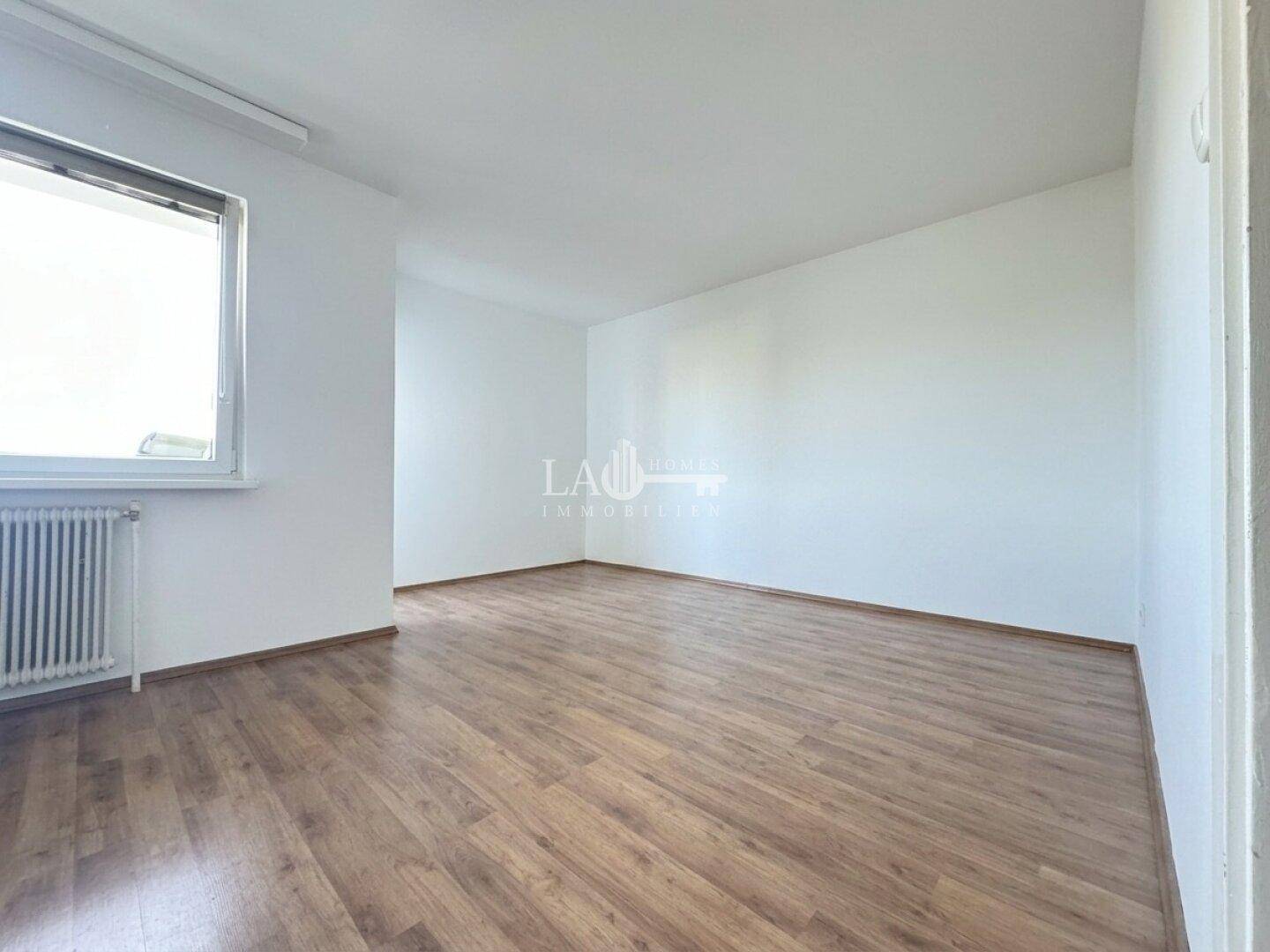Heller, leerer Raum mit Laminatboden, Fenster und viel Tageslicht.