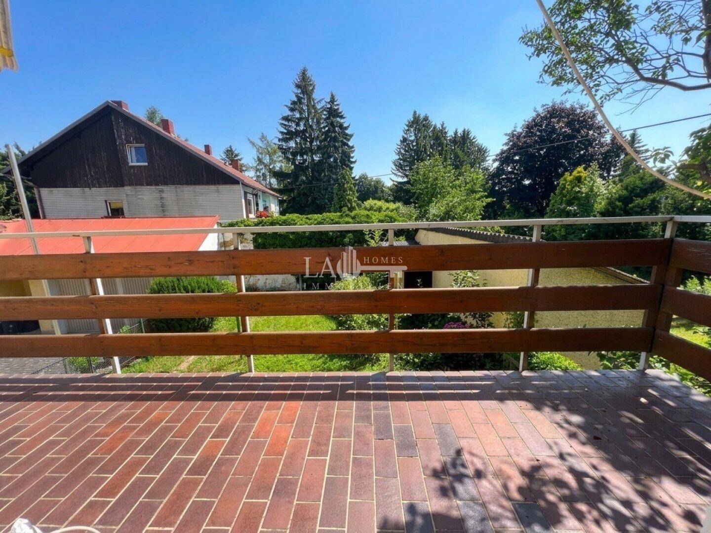 Geräumige Terrasse mit Ziegelpflaster und Holzgeländer, bietet einen Blick auf umliegende Häuser und viel Grün.