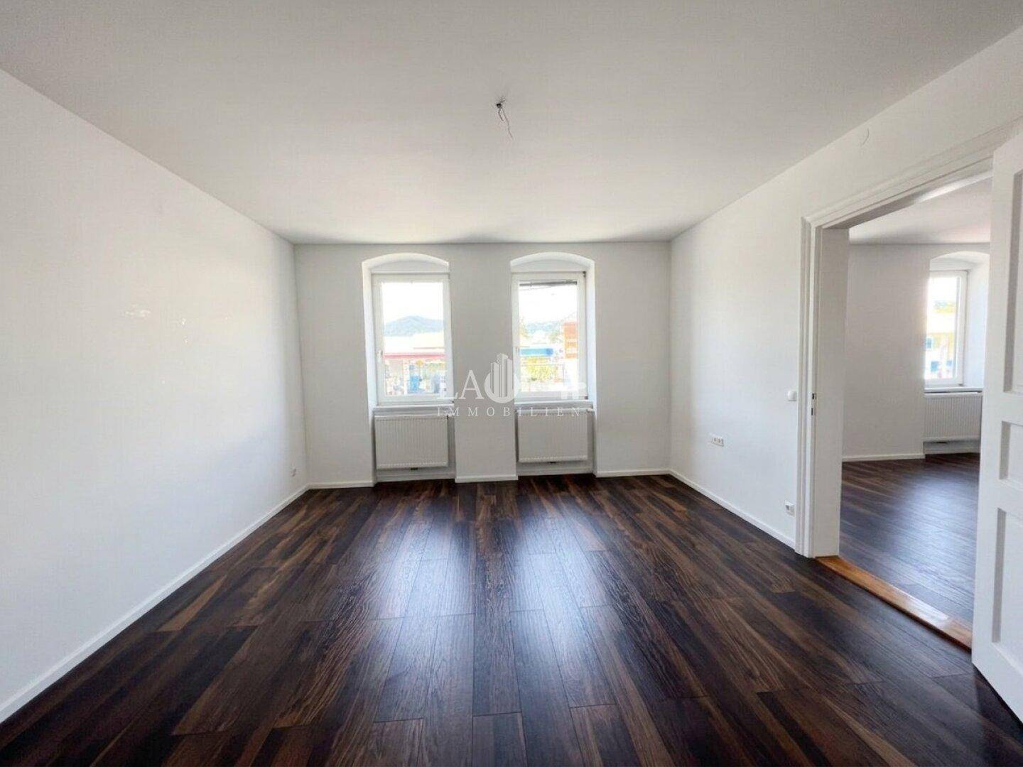 Geräumiges Wohnzimmer mit dunklem Laminatboden und weißen Wänden, bietet durch zwei Fenster viel Tageslicht.