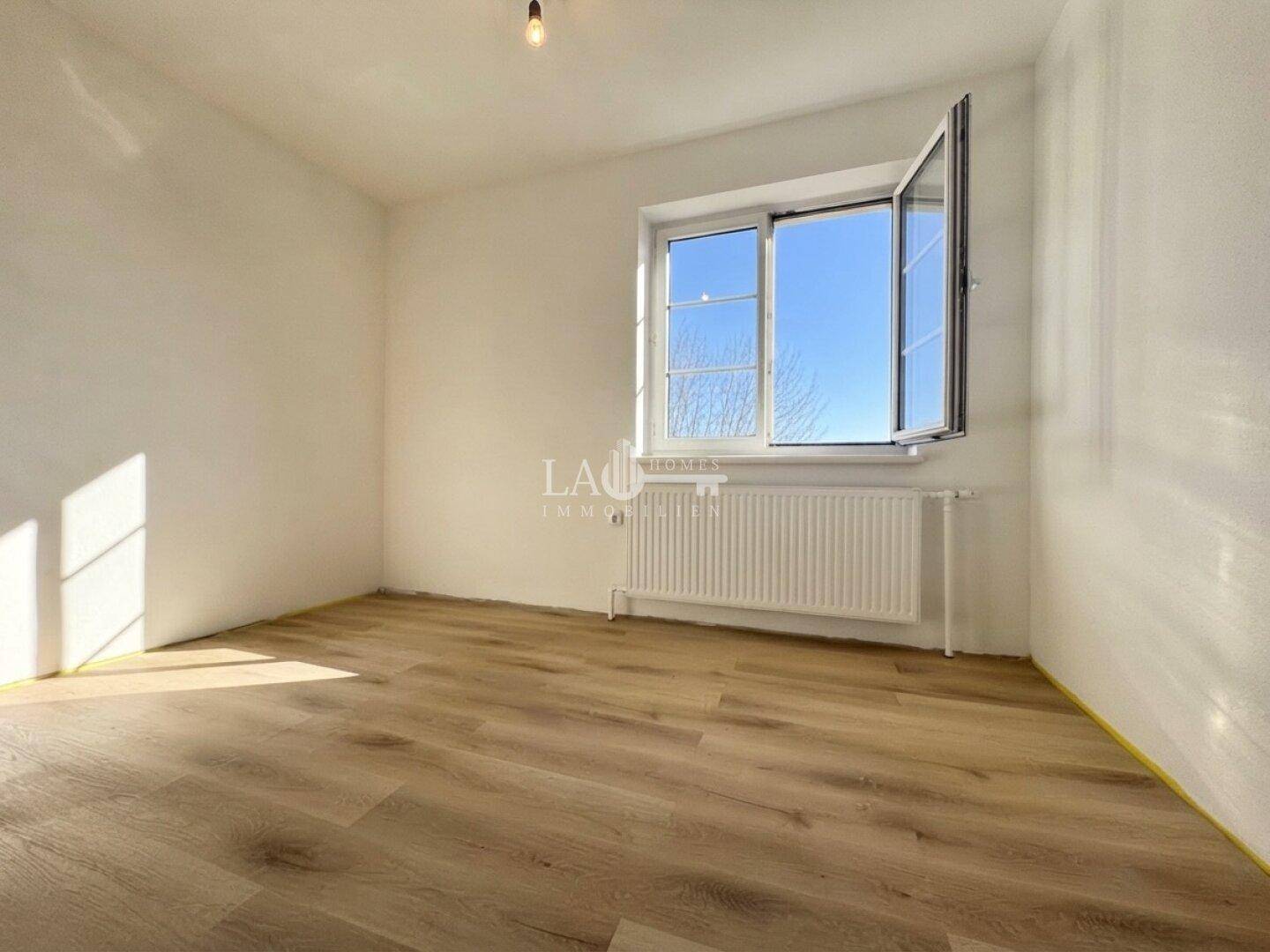 Lichtdurchfluteter, leerer Raum mit einem Fenster, Laminatboden und einem Ausblick ins Grüne.