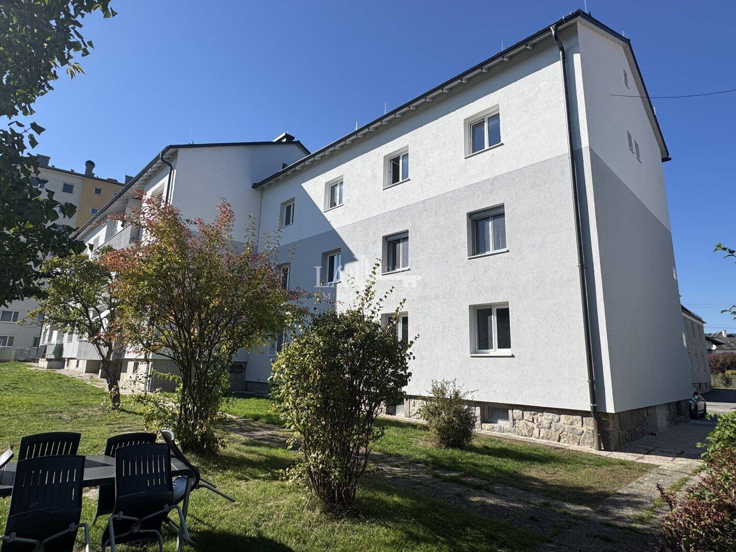 Rückseite des Wohngebäudes mit gepflegtem Rasen, Bäumen und einer kleinen Sitzgelegenheit im Freien.