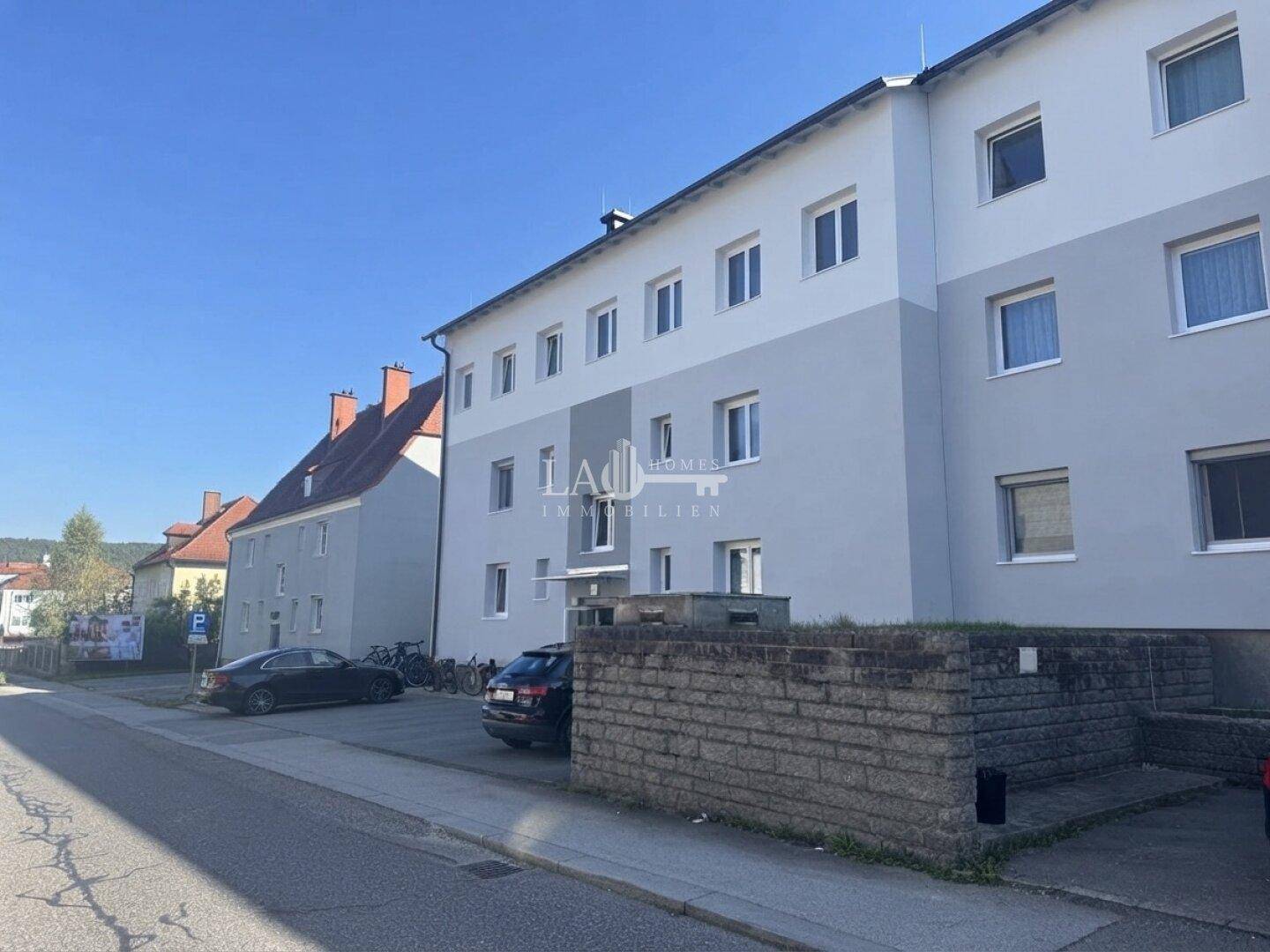 Ansicht des Wohngebäudes von der Straße mit geparkten Autos und einer niedrigen Steinmauer im Vordergrund.