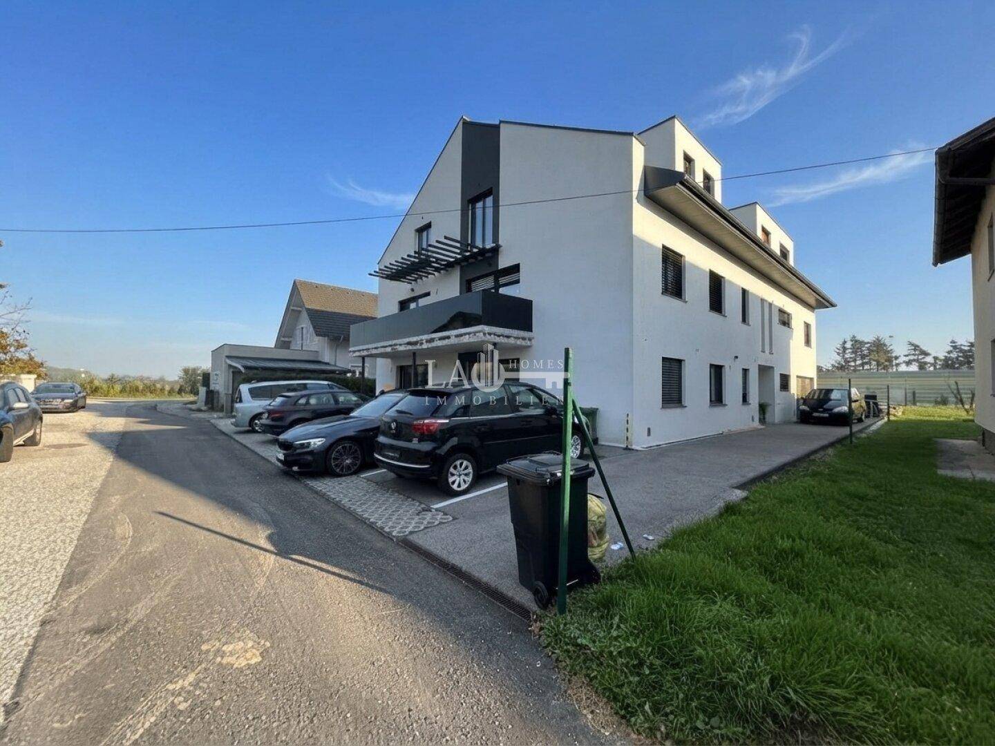 Modernes Mehrfamilienhaus mit weißer Fassade, Parkplätzen und grüner Umgebung an einem sonnigen Tag.