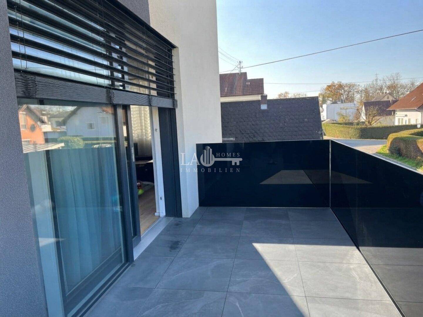 Moderner Balkon mit grauen Fliesen und schwarzer Glasbrüstung, bietet einen Blick auf die Umgebung.