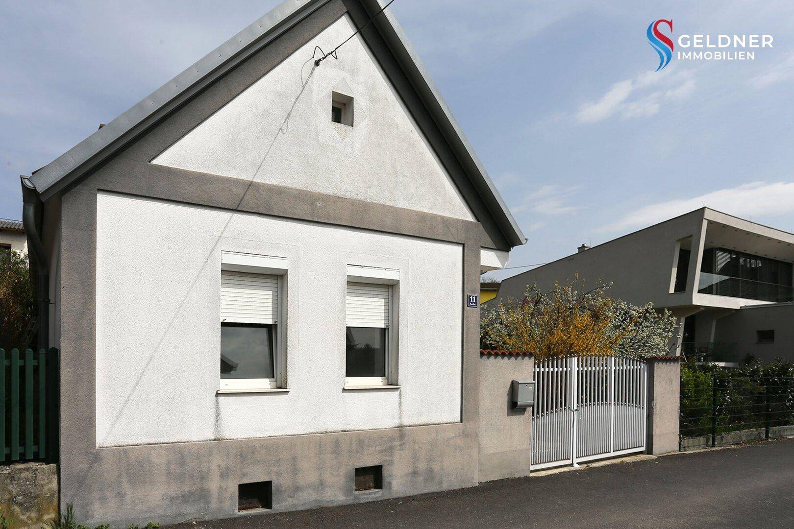 Vorderansicht des Hauses mit weißer Fassade, Rollläden und einem modernen Nachbargebäude.