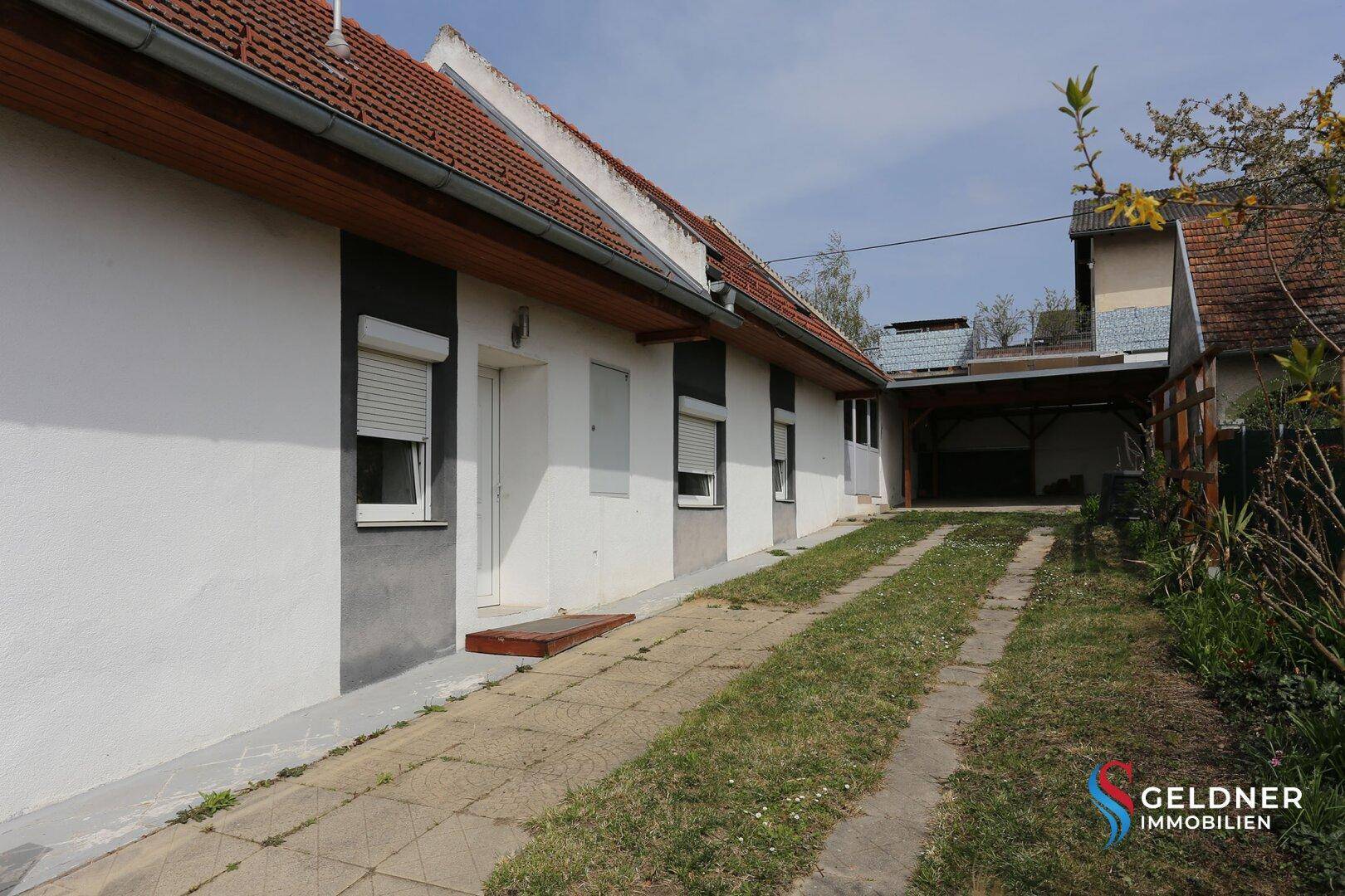 Seitenansicht des Hauses mit Zufahrt, Rasenfläche und einem Carport im Hintergrund.
