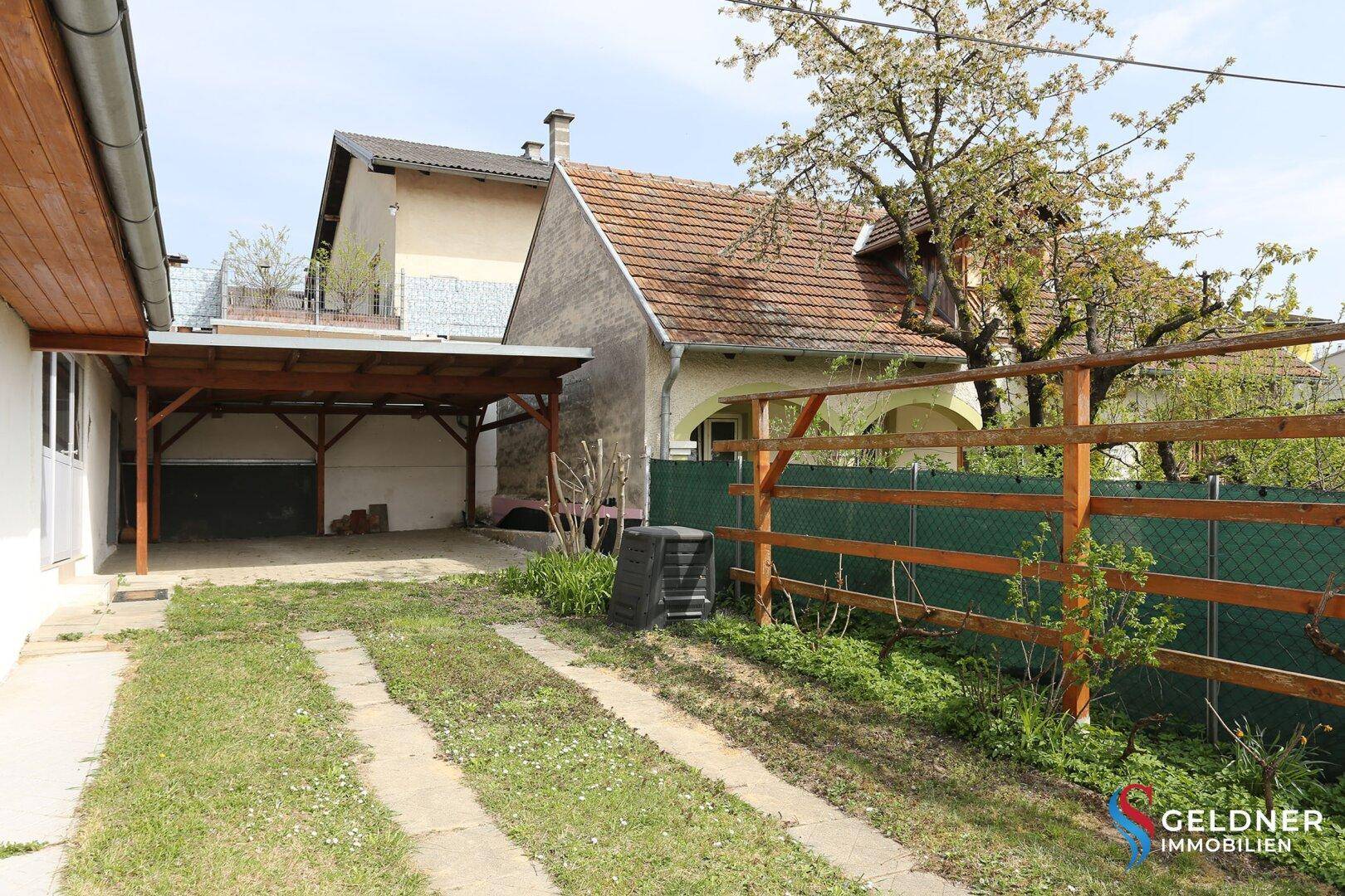 Hinterhofbereich mit Carport, Rasenfläche und einem Komposter.