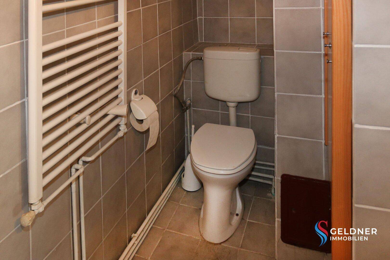 Toilette mit Heizkörper, WC und grauen Fliesen an Wänden und Boden.