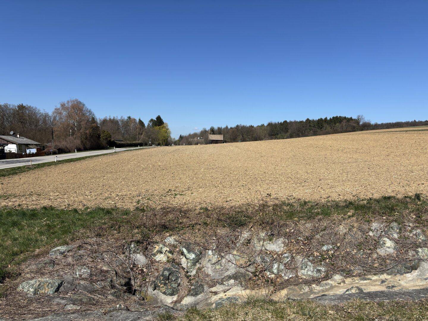 Ackerland mit einer Straße, Häusern in der Ferne und einer Steinmauer im Vordergrund unter blauem Himmel.
