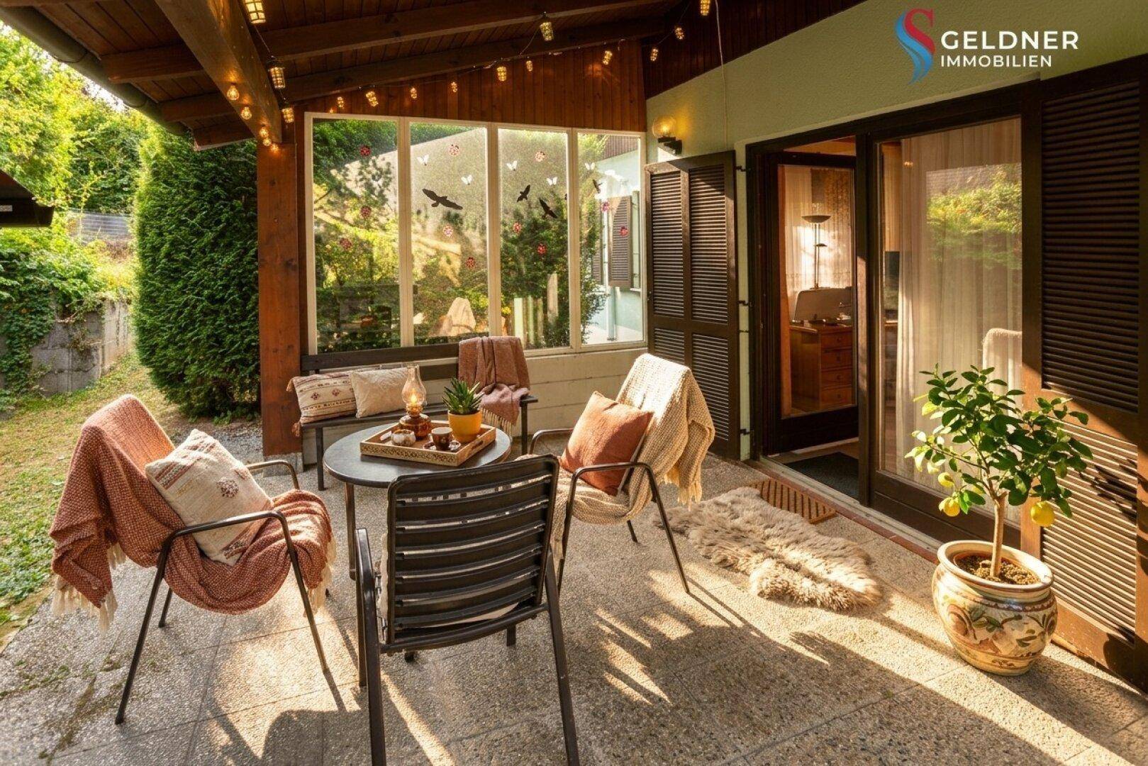 Sonnige Terrasse mit Sitzgelegenheiten, umgeben von Pflanzen und mit Zugang zum Innenbereich.