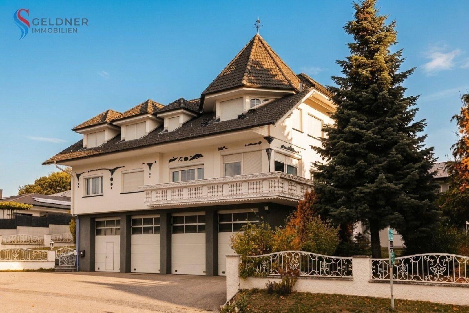 Vorderansicht des Hauses mit Turmdach, Balkon und drei Garagentoren, umgeben von hohen Bäumen.