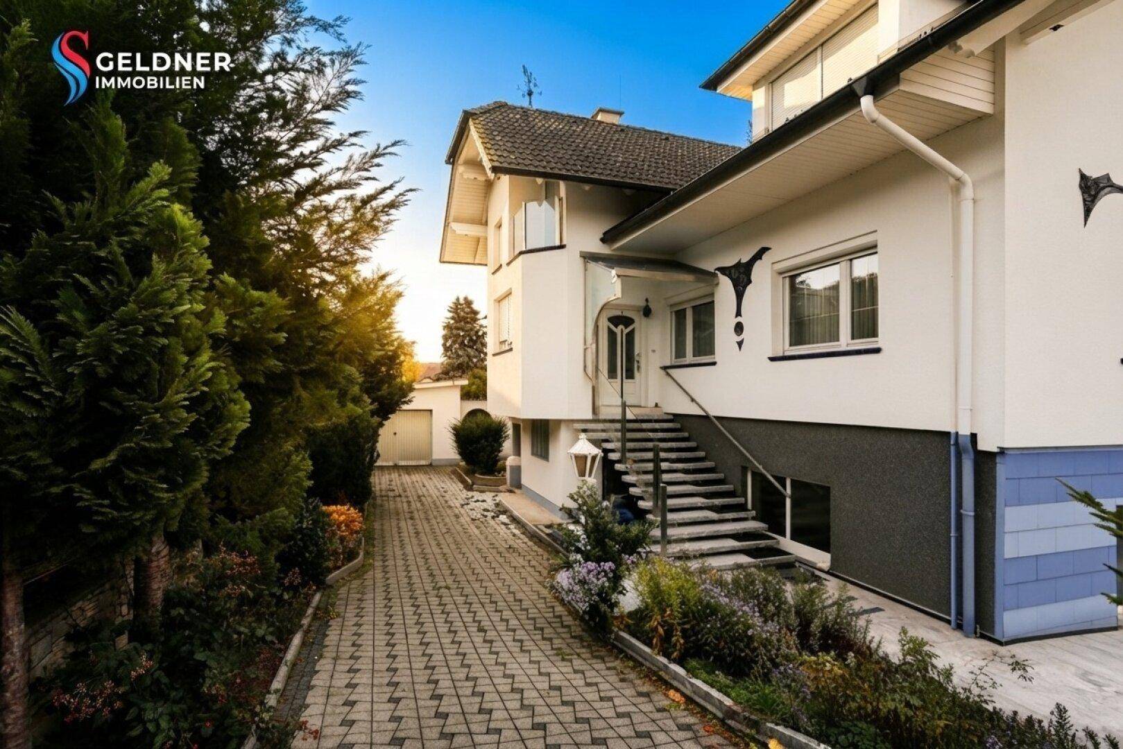 Gepflasterte Auffahrt zum Hauseingang mit Treppe und üppiger Bepflanzung im Vorgarten.