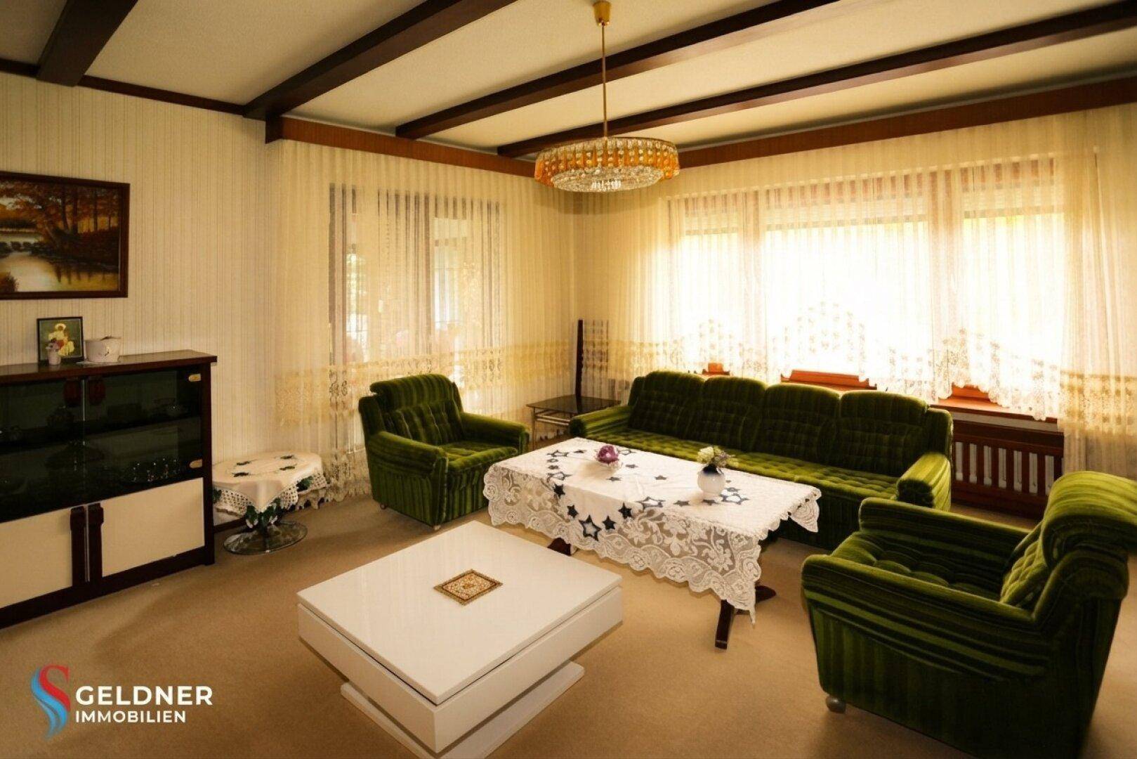 Gemütliches Wohnzimmer mit grünen Samtsofas, Holzbalkendecke und großem Fenster.
