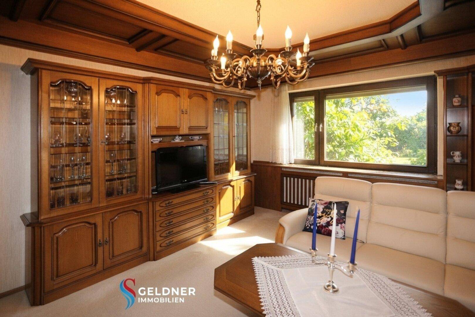 Wohnzimmer mit Holztäfelung, Ledersofa und Kronleuchter, mit Blick ins Grüne.
