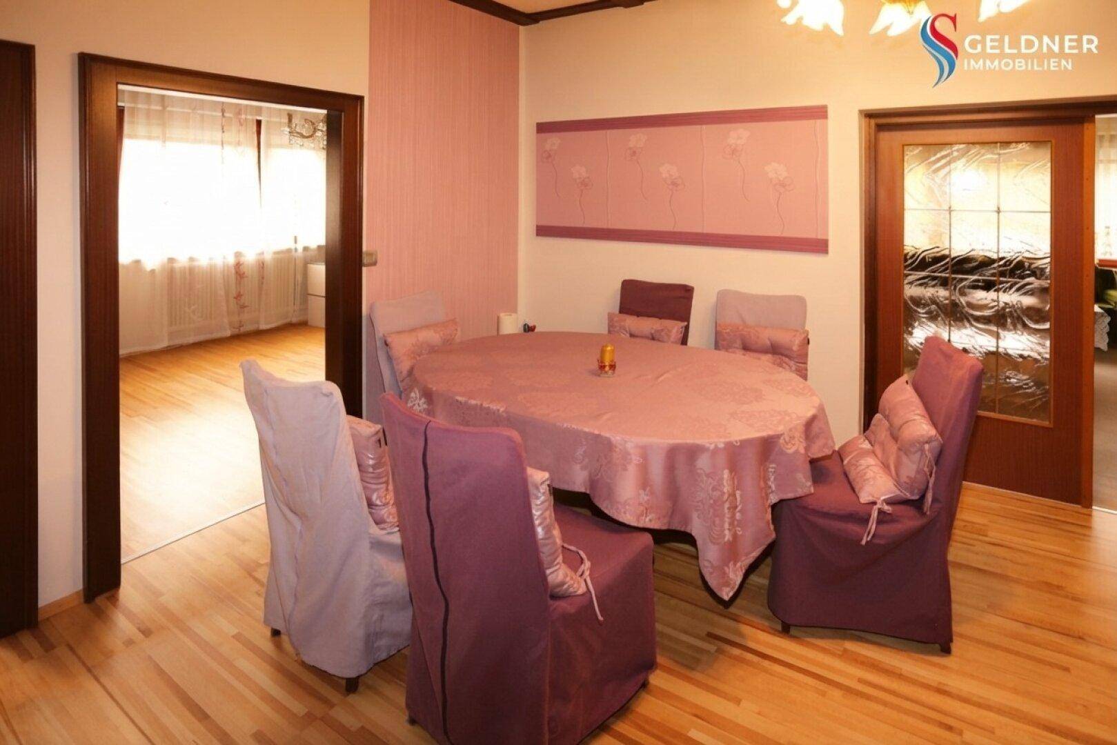Esszimmer mit Holzboden, großem Esstisch und rosa Wandgestaltung, einladend und hell.