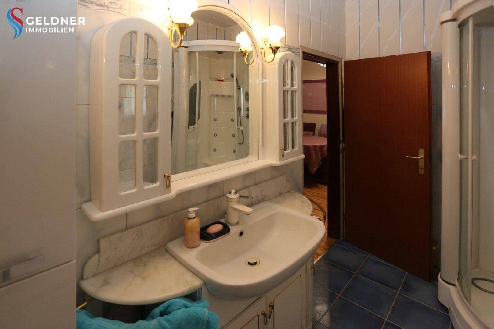 Badezimmer mit Waschtisch, Spiegelschrank und Duschkabine, durch eine offene Tür sichtbar.