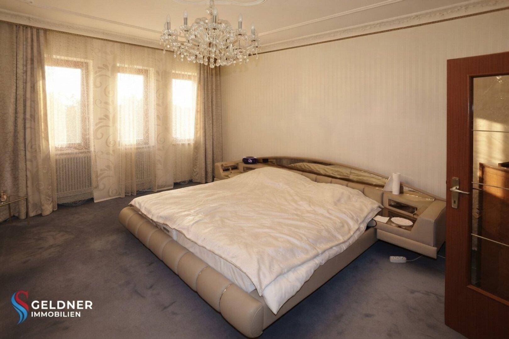 Schlafzimmer mit großem Bett, Kronleuchter und Fenstern mit Vorhängen, in einem klassischen Stil.