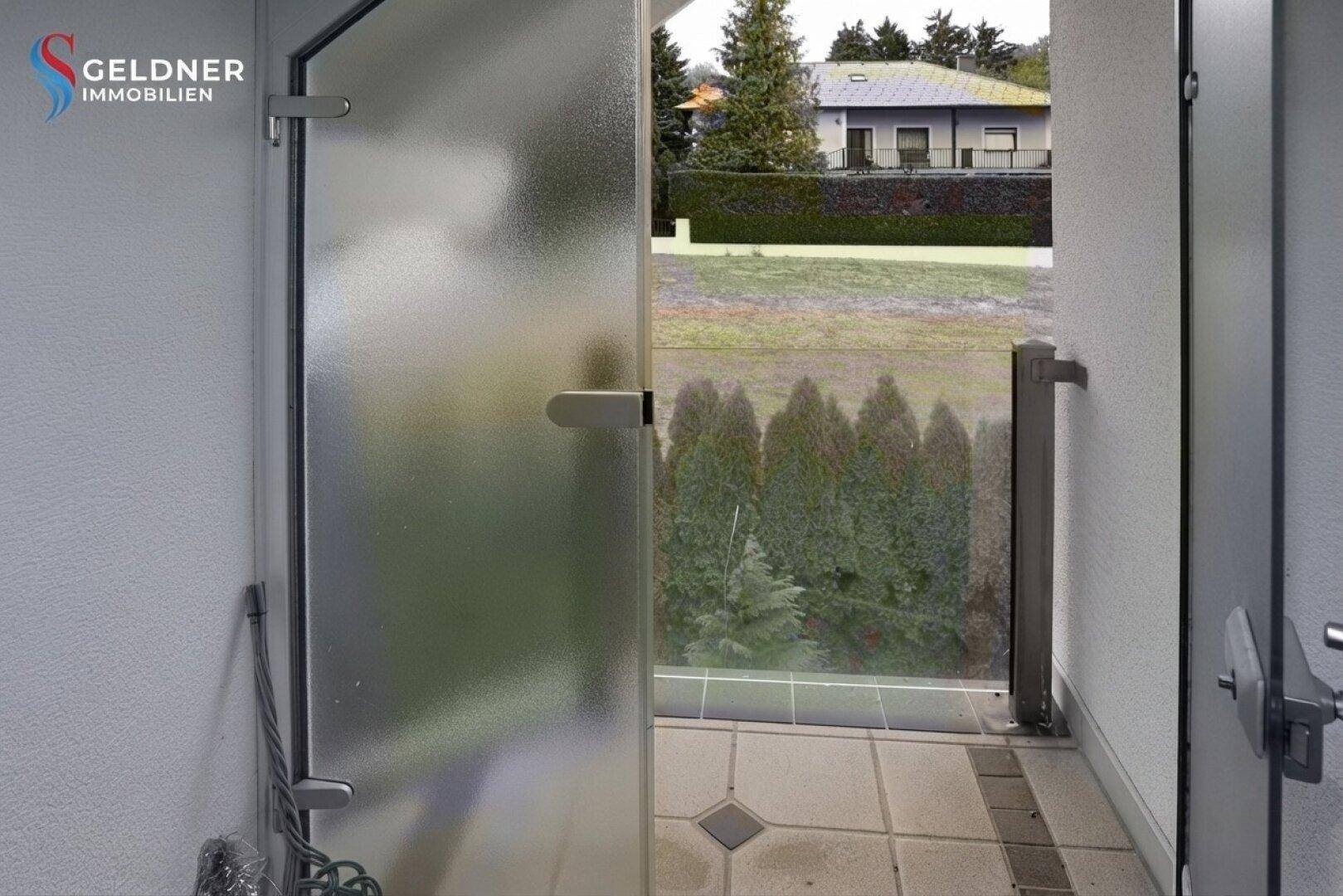 Blick von einer verglasten Terrasse auf den Garten und die umliegende Nachbarschaft mit viel Grün.